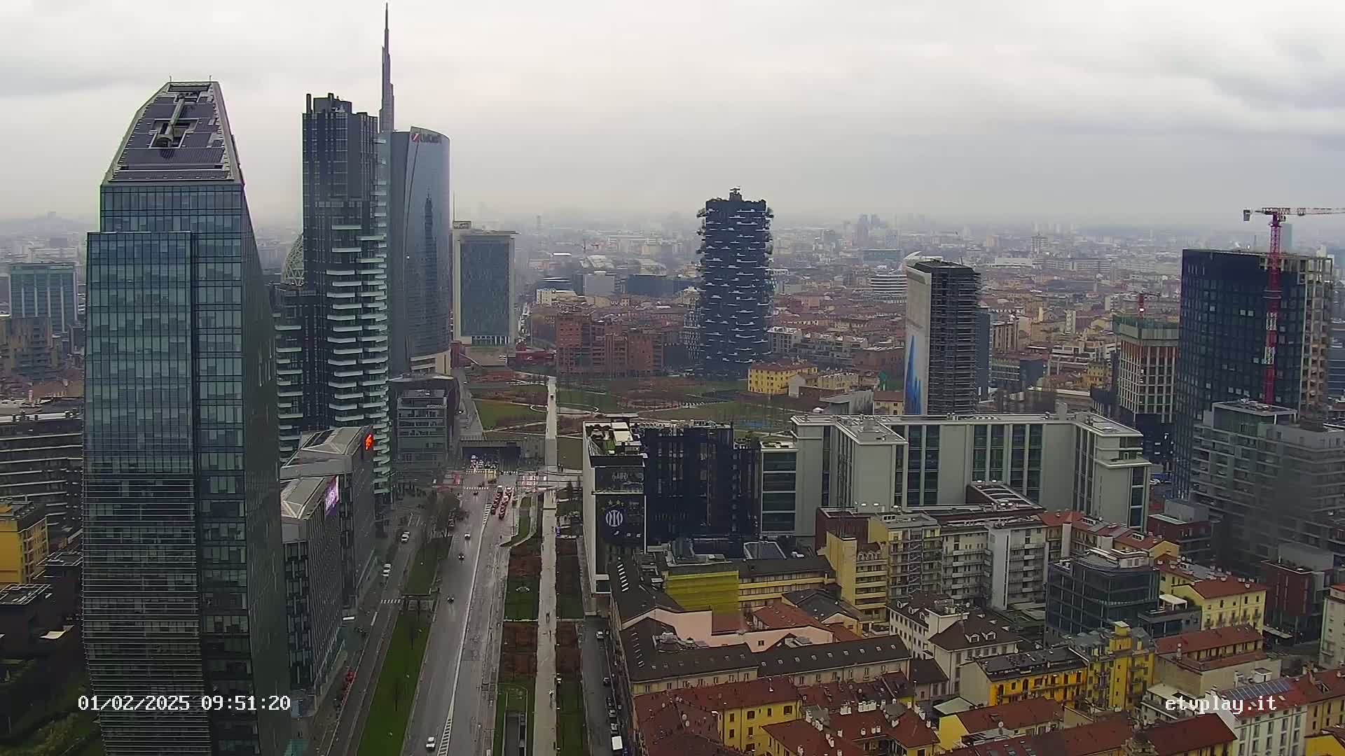 An aerial view of a city's skyline on an overcast day shows several modern skyscrapers and surrounding buildings, with a long road visible below.