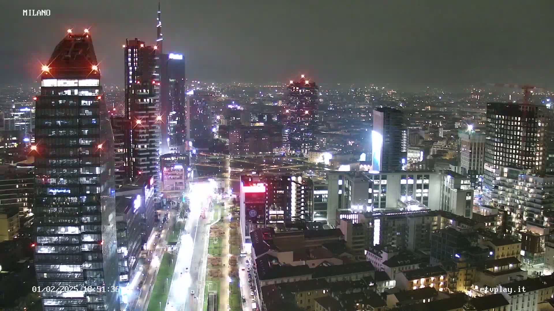 A nighttime, high-angle view of a city skyline with many skyscrapers, lit up streets below, and a slightly hazy atmosphere.