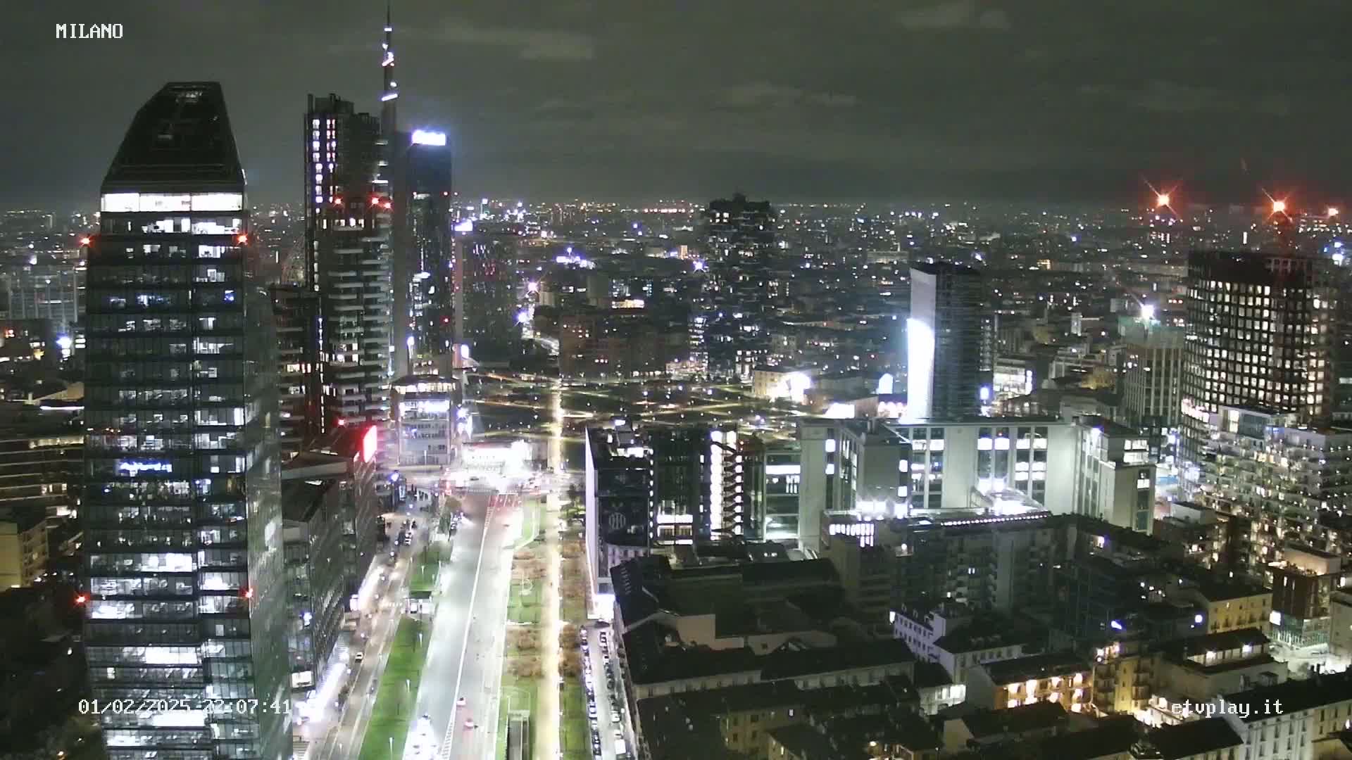 A nighttime aerial view of a city, showing numerous illuminated buildings and streets, under a dark, overcast sky.