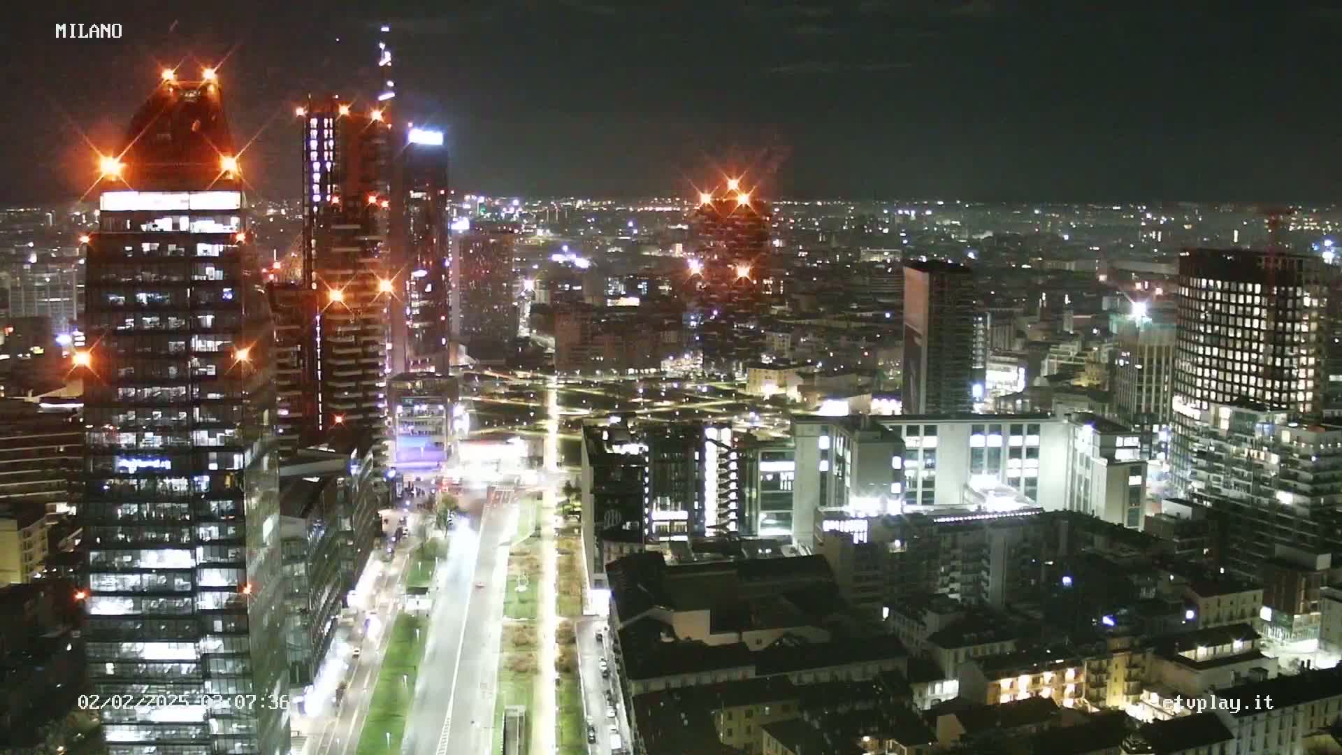 A nighttime aerial view of a city shows numerous illuminated buildings, roads, and a long, landscaped area stretching towards the distance under a clear sky.