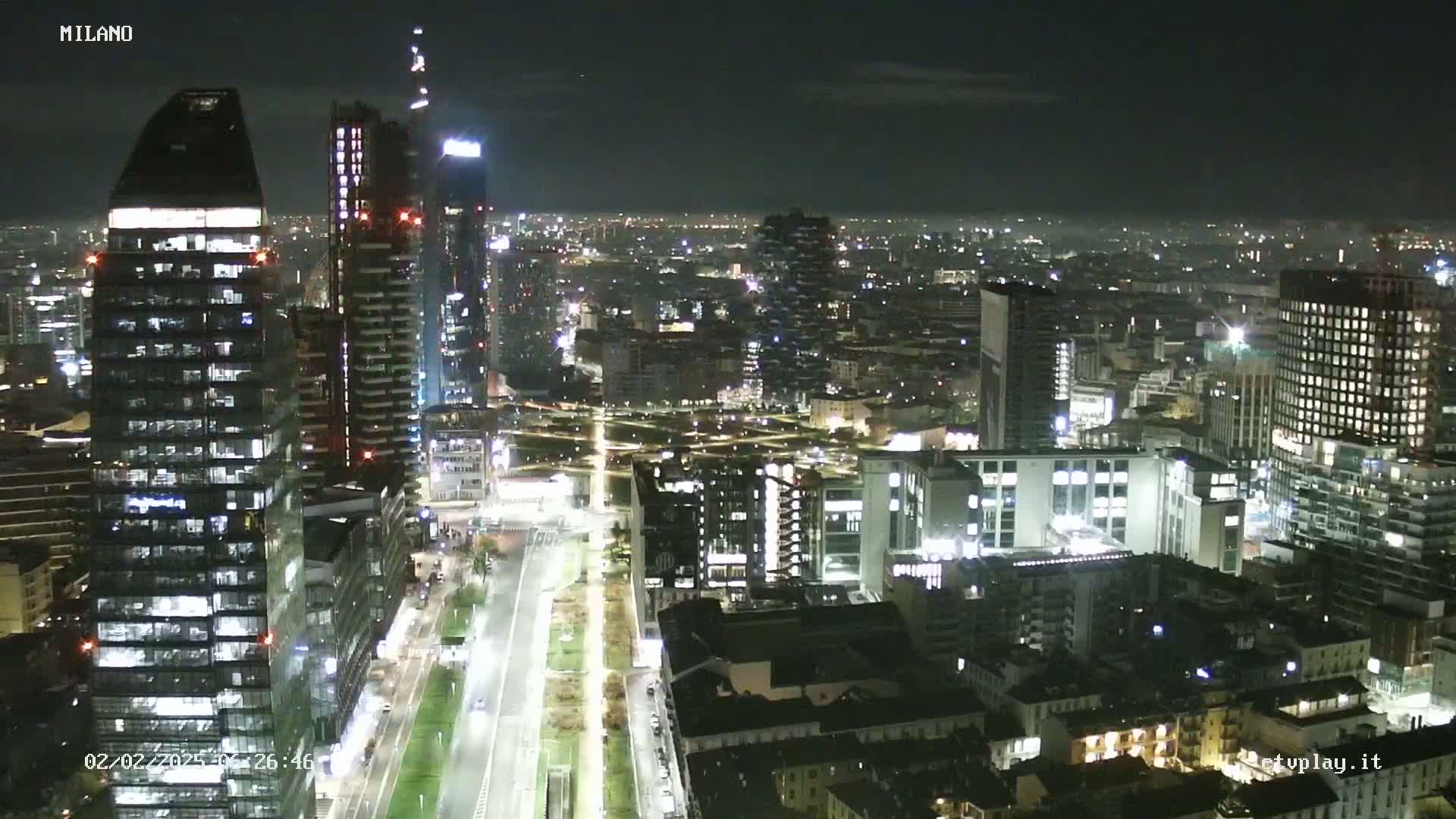 A nighttime, high-angle view of a cityscape showing numerous illuminated buildings, streets, and a long, tree-lined avenue.