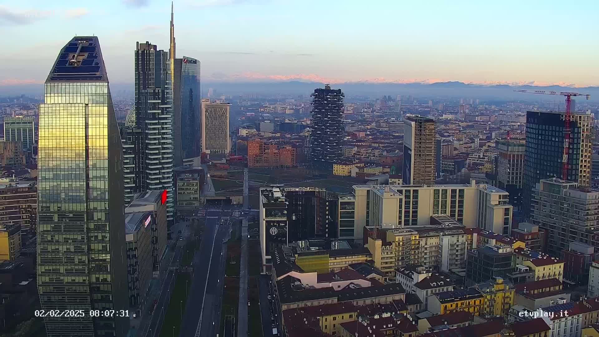 A hazy sunrise illuminates a cityscape of numerous skyscrapers and buildings, with distant mountains visible on the horizon.