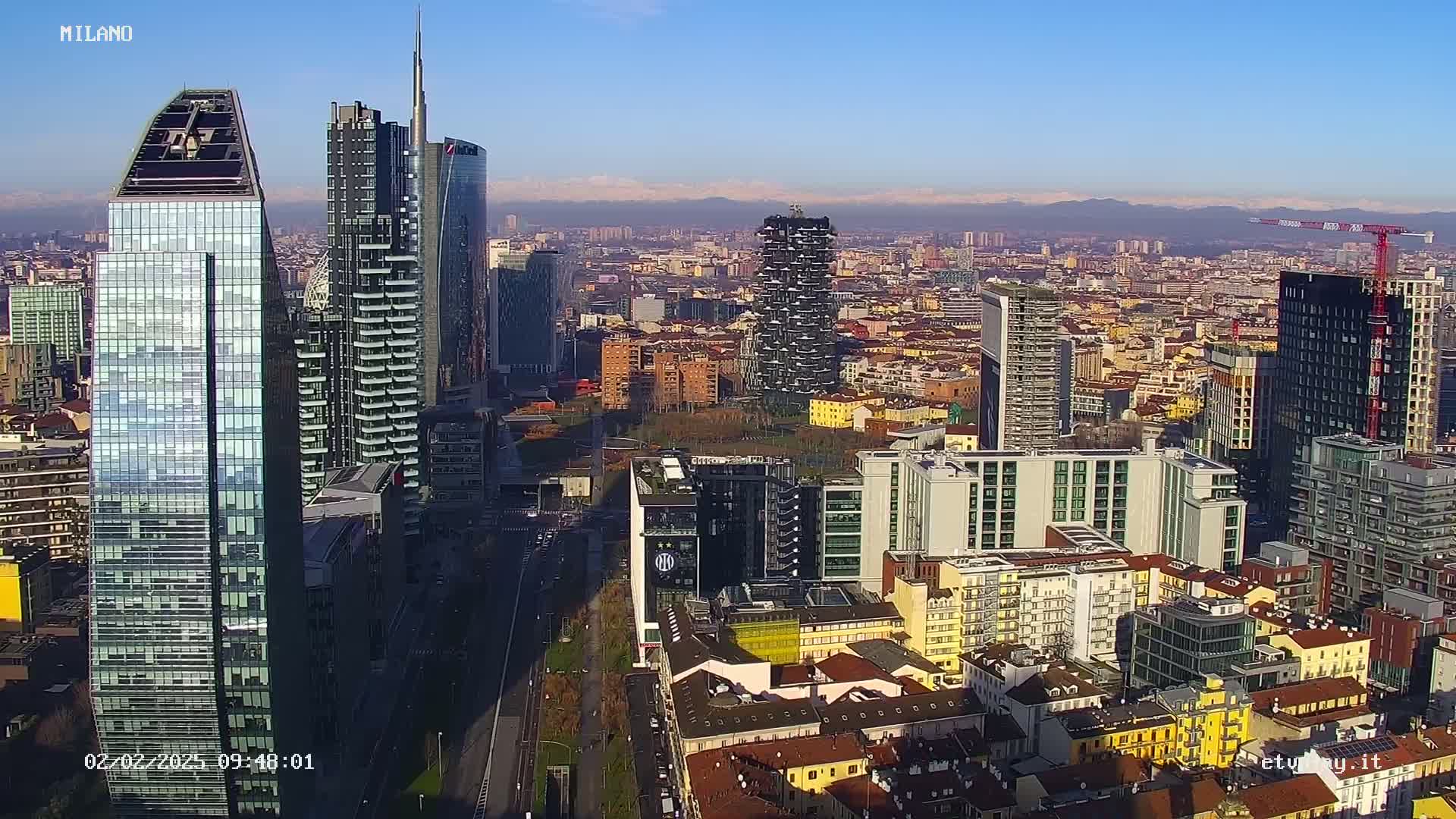 An aerial view of a dense cityscape with numerous skyscrapers under a clear, sunny sky, mountains visible in the distance.