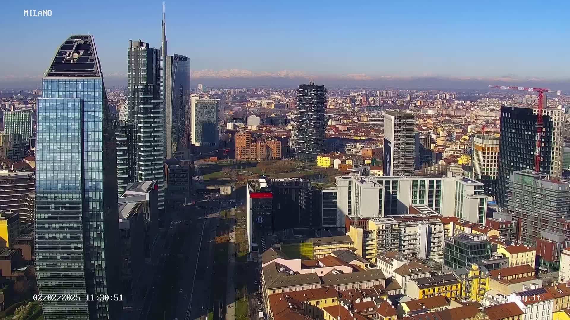 An aerial view of a city skyline with many skyscrapers on a clear, sunny day, with distant mountains visible in the background.