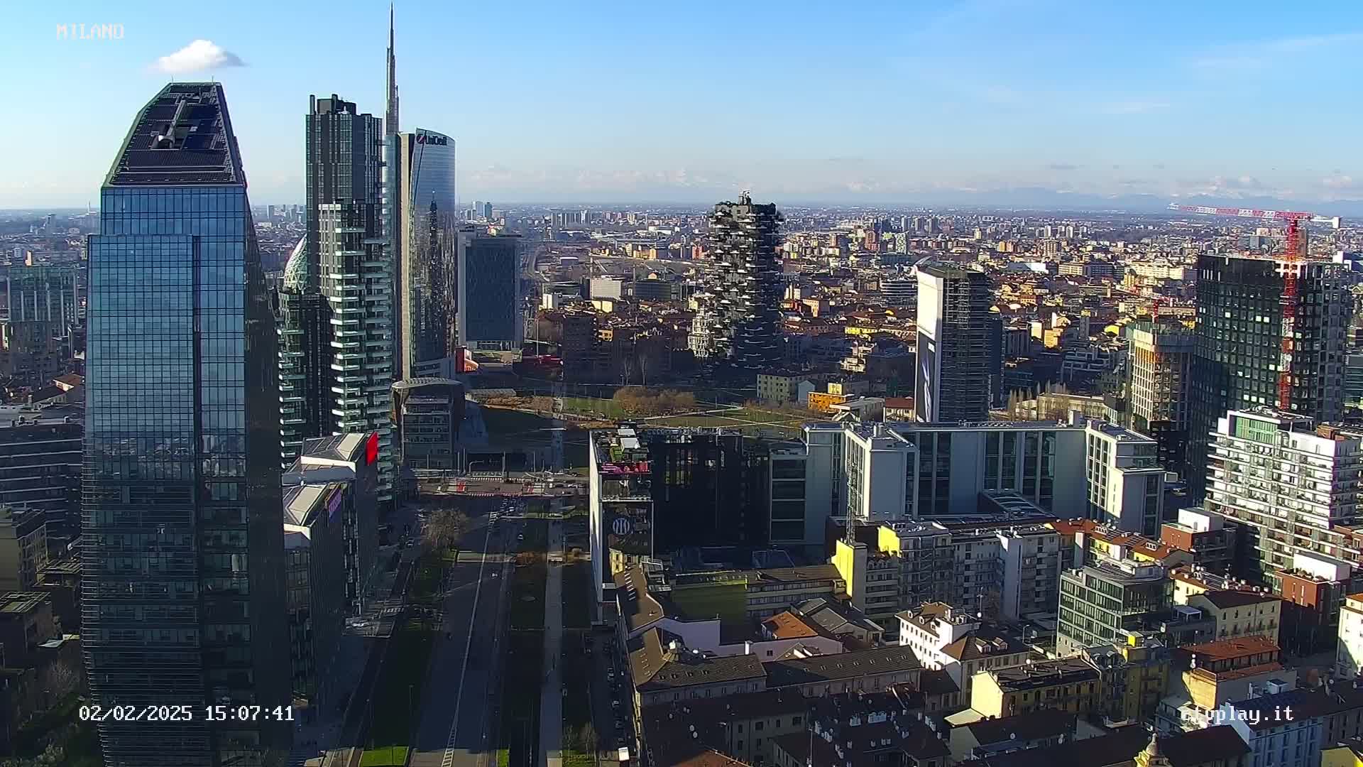 An aerial view of a city skyline on a clear, sunny day, showing a cluster of modern skyscrapers surrounded by a mix of taller and shorter buildings.