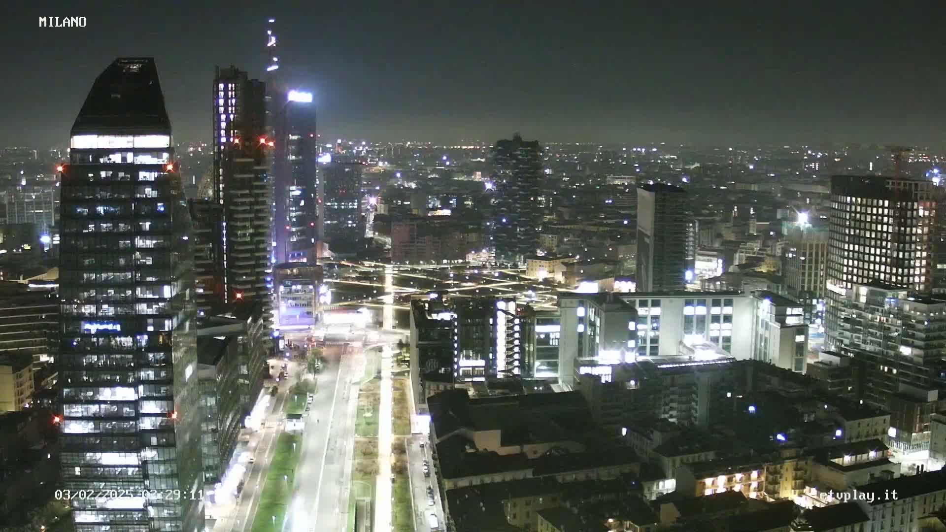 A nighttime aerial view of a city, showing brightly lit skyscrapers and streets, under a clear, dark sky.