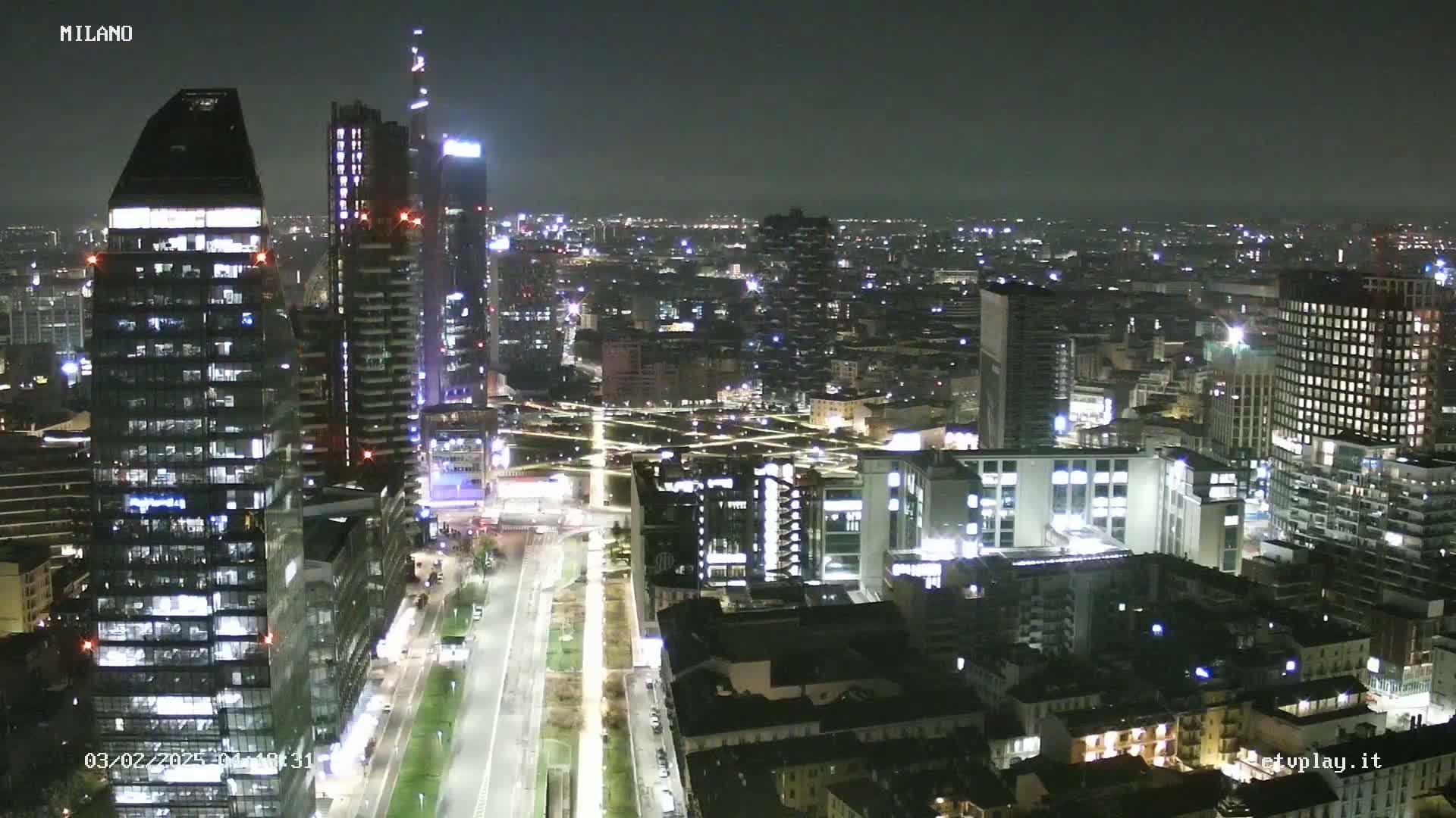 A nighttime aerial view of a city with numerous illuminated buildings, streets, and a long, brightly lit road stretching into the distance.