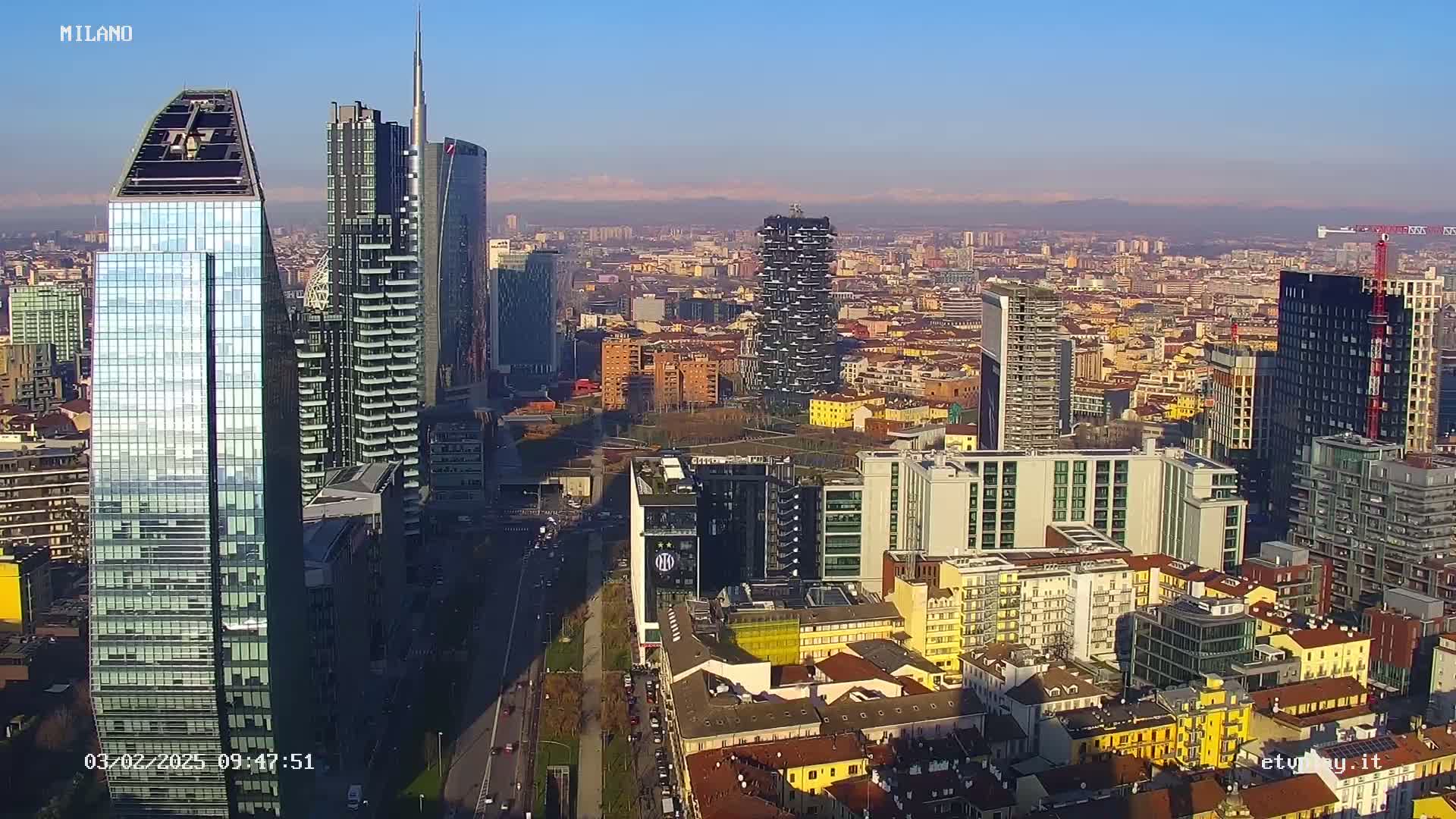 A high-angle, long shot of a cityscape shows many modern skyscrapers and other buildings on a clear, sunny day with distant mountains visible in the background.