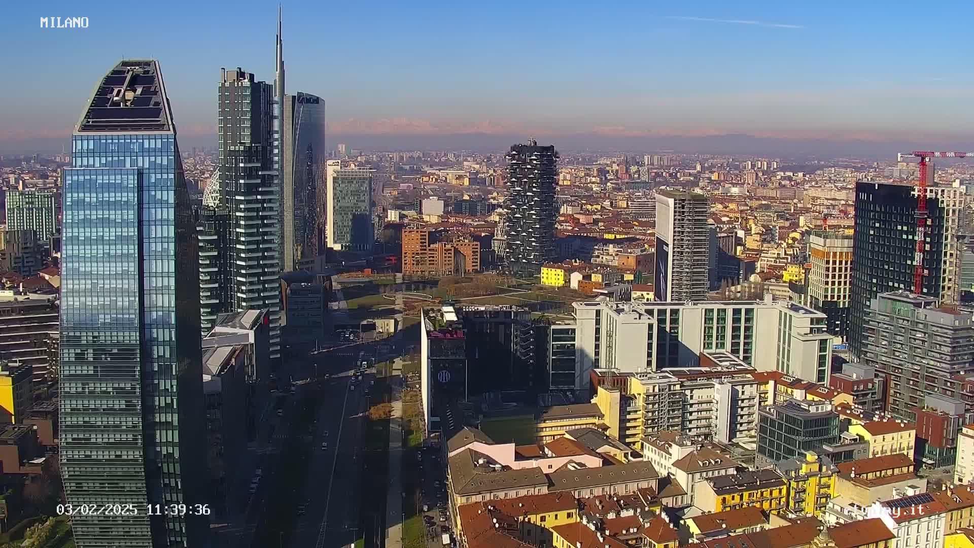 A high-angle, long shot of a dense cityscape of modern and older buildings under a clear, sunny sky with distant mountains visible on the horizon.