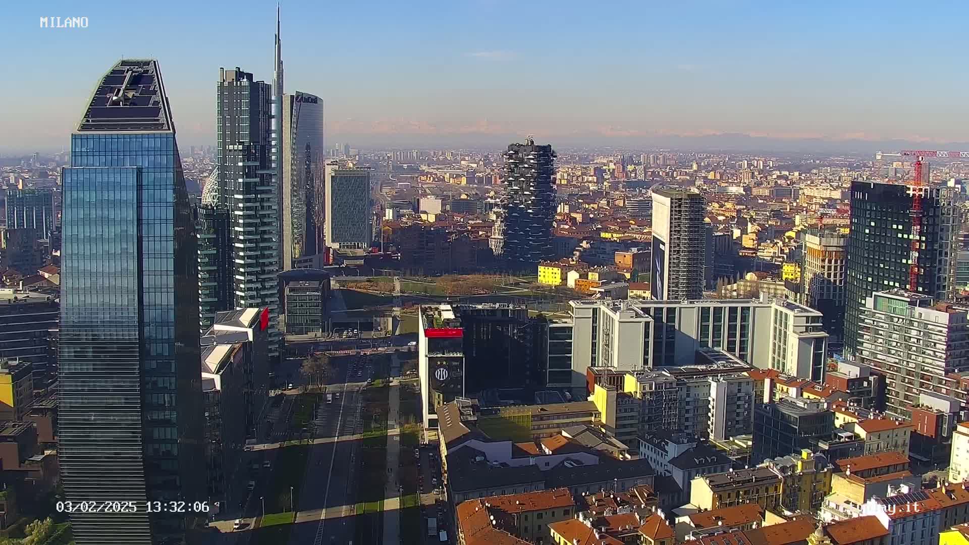 A high-angle, long shot of a dense cityscape on a sunny day, featuring numerous skyscrapers and other buildings, with distant mountains visible on the horizon.