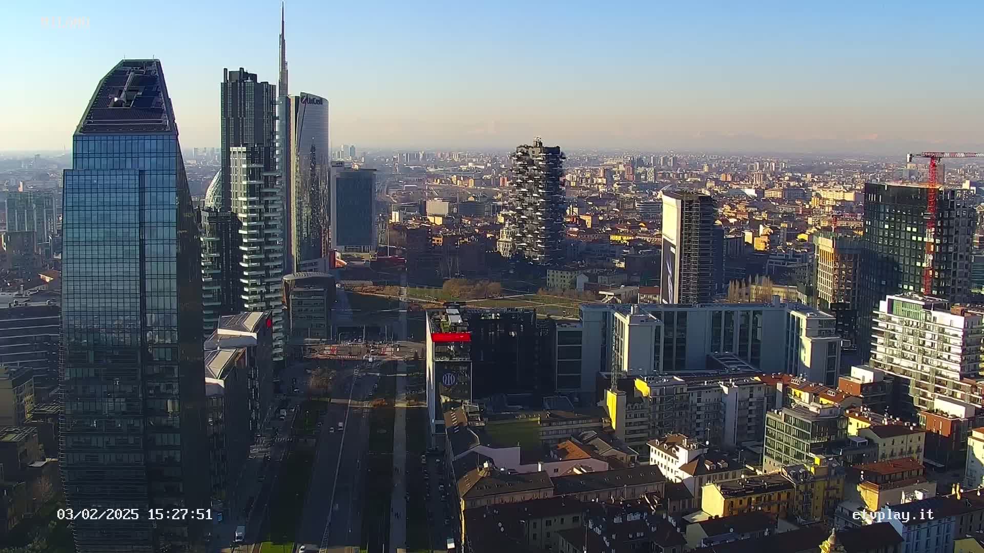 An aerial view of a city skyline on a clear, sunny day, showing numerous skyscrapers and surrounding buildings.