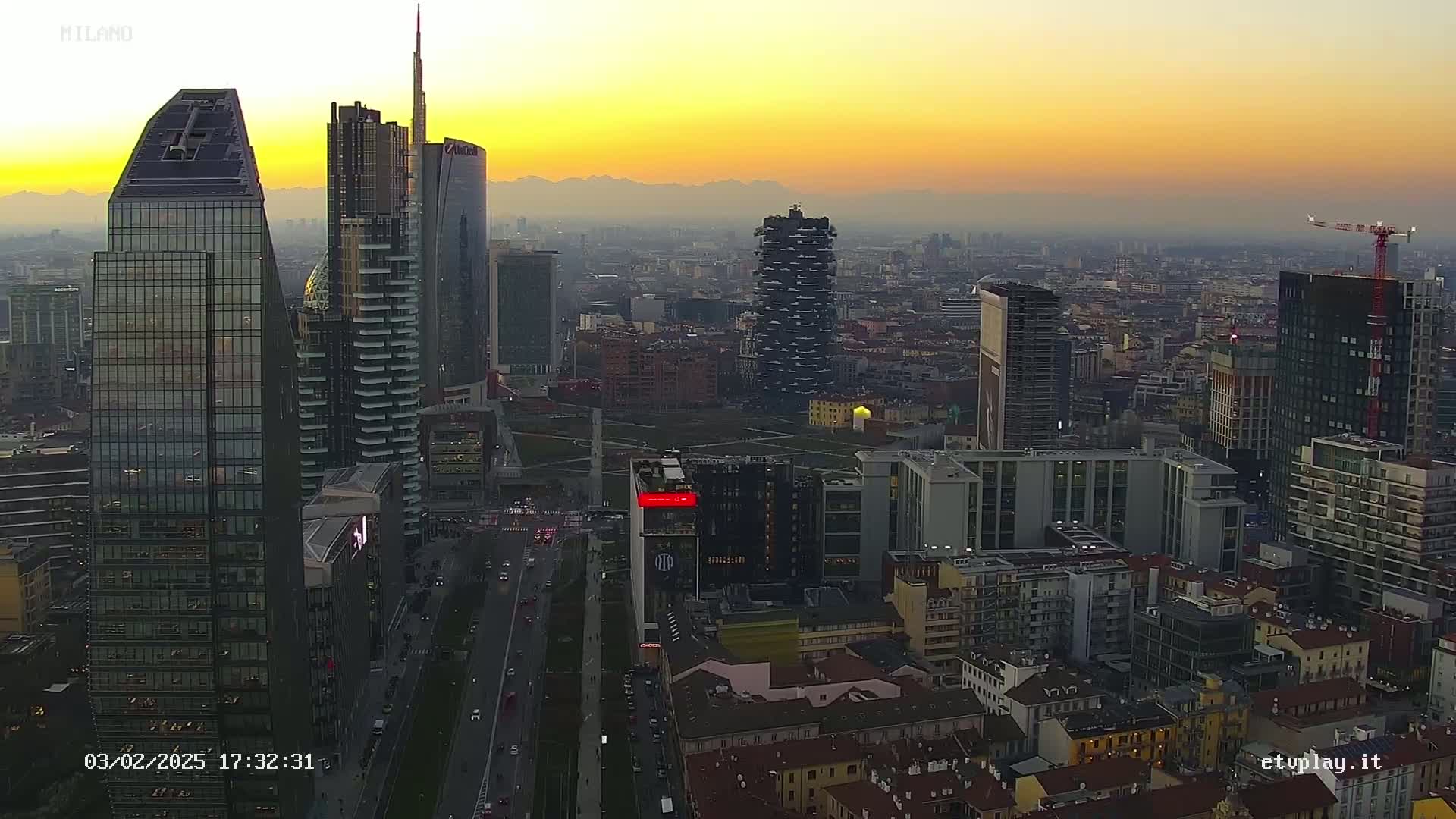 An aerial view of a city skyline at sunset, showing numerous skyscrapers and buildings, under hazy atmospheric conditions with distant mountains visible.