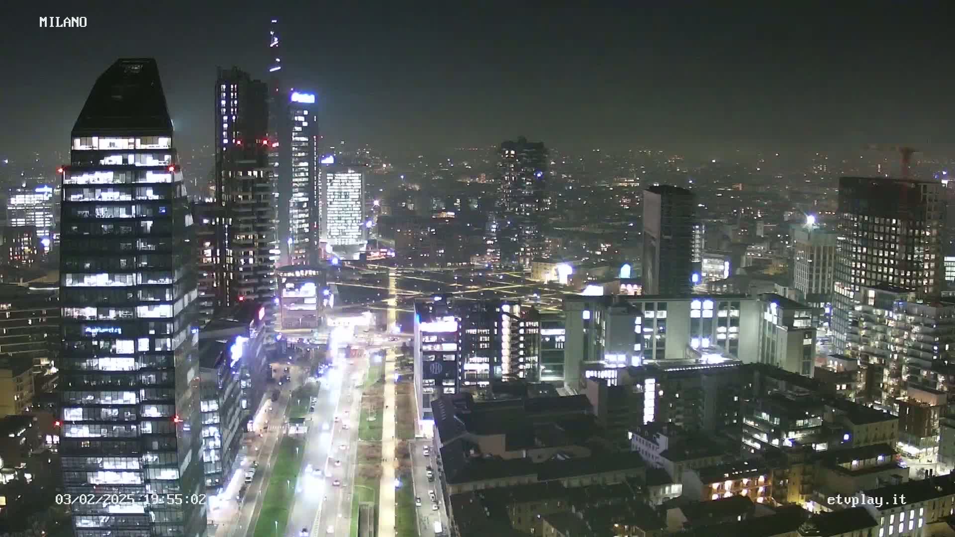 A nighttime aerial view of a city skyline showing numerous illuminated skyscrapers and streets with moving traffic under a hazy sky.