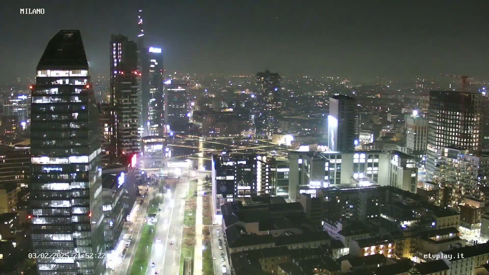 A nighttime aerial view of a city skyline, showing numerous illuminated skyscrapers and buildings, with streets and roadways visible below.