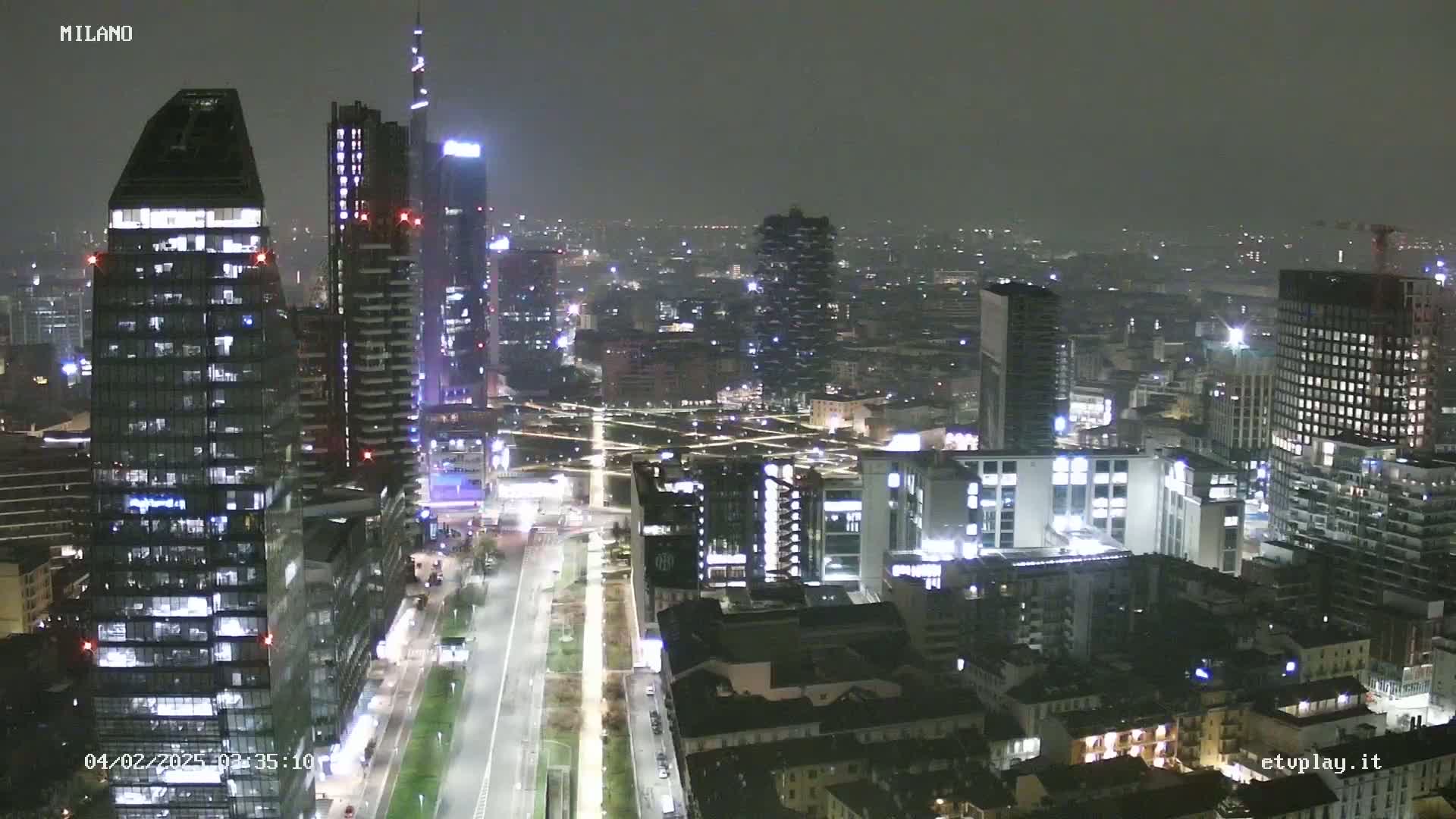 A nighttime aerial view of a city, possibly Milan, showing numerous illuminated skyscrapers and buildings under a hazy sky.