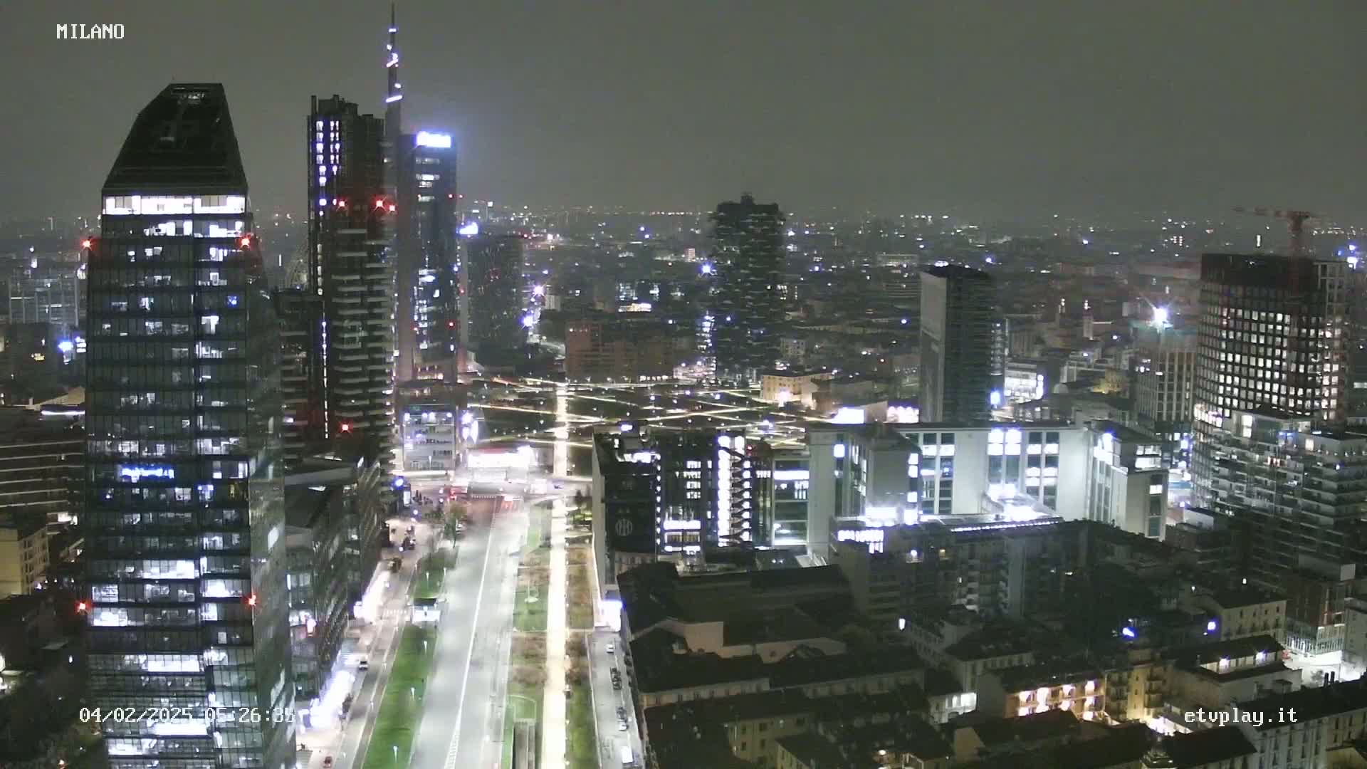 A nighttime aerial view of a city's skyline, showing numerous illuminated skyscrapers and buildings under a dark, overcast sky.