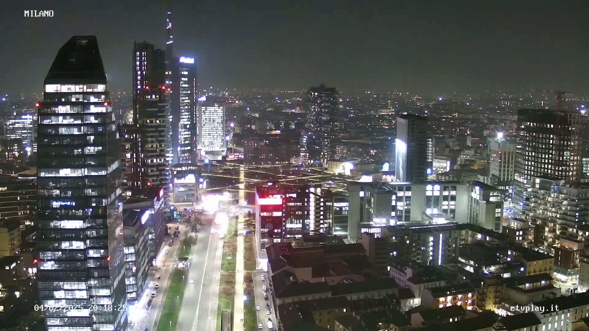 A nighttime aerial view of a city's skyline, showing numerous illuminated skyscrapers and streets with sparse traffic under a dark sky.