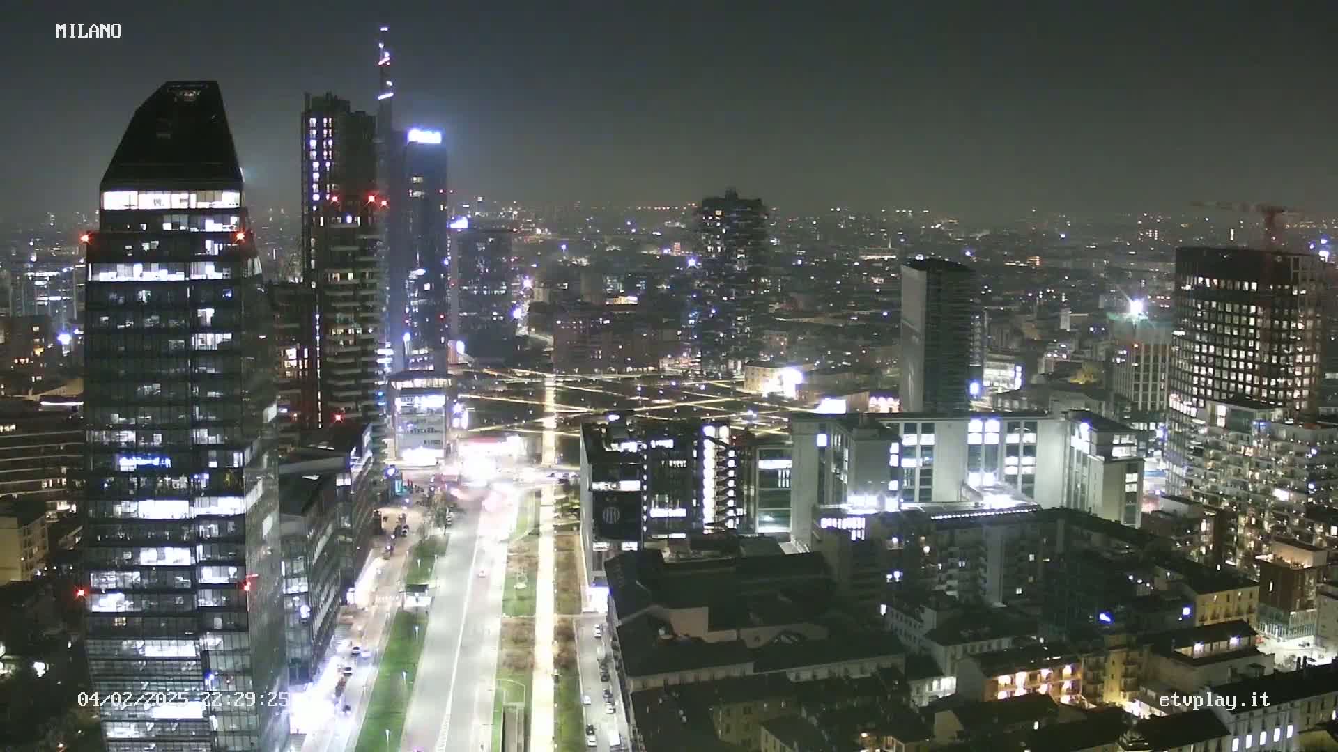 A nighttime aerial view of a city skyline with many illuminated skyscrapers and buildings under a hazy sky.