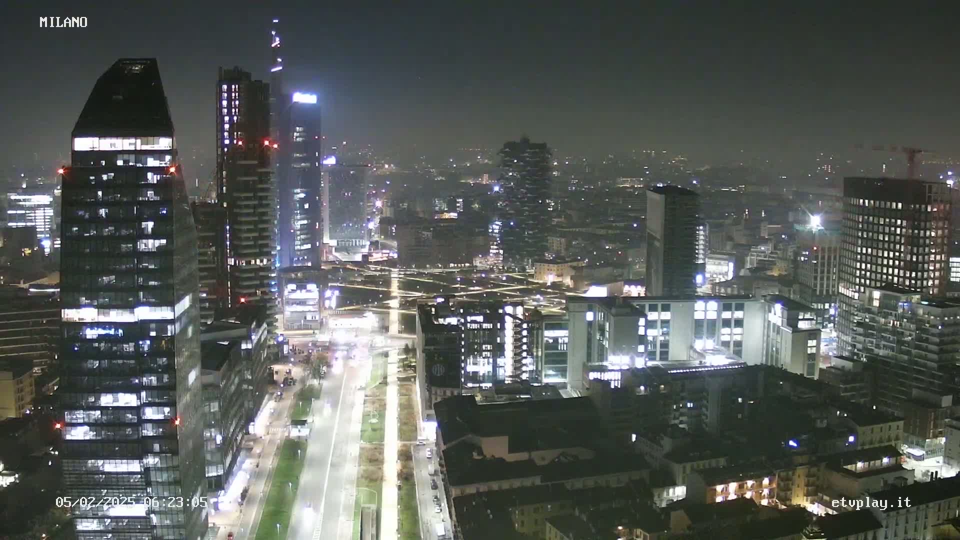 A nighttime aerial view of a city skyline with numerous illuminated skyscrapers and buildings, under a hazy sky.