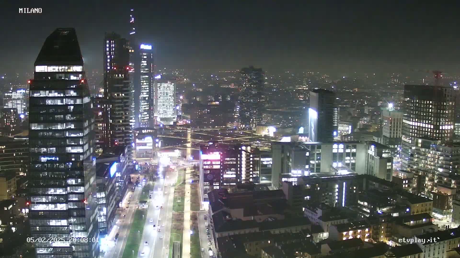 A nighttime aerial view of a city skyline showing numerous skyscrapers and brightly lit streets under hazy conditions.