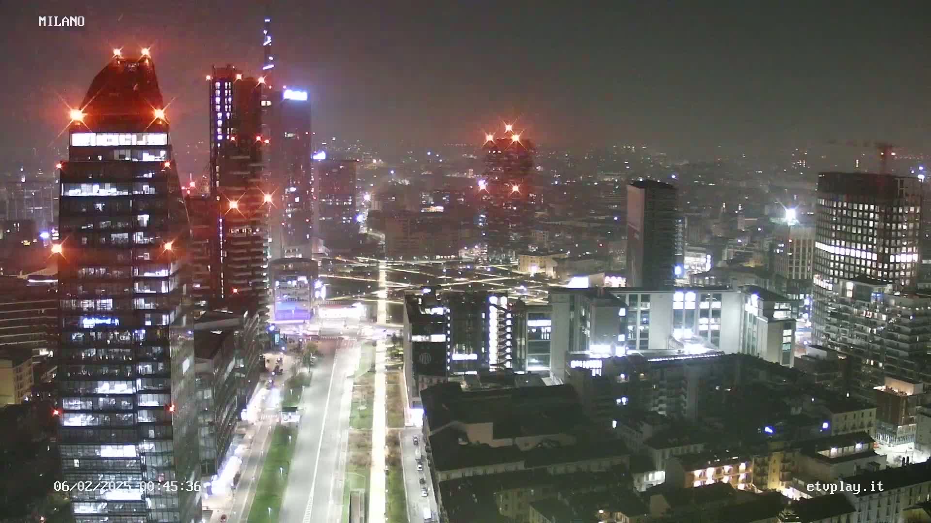 A nighttime, hazy view of a city skyline, showing numerous illuminated skyscrapers and buildings, with streets and roads visible below.