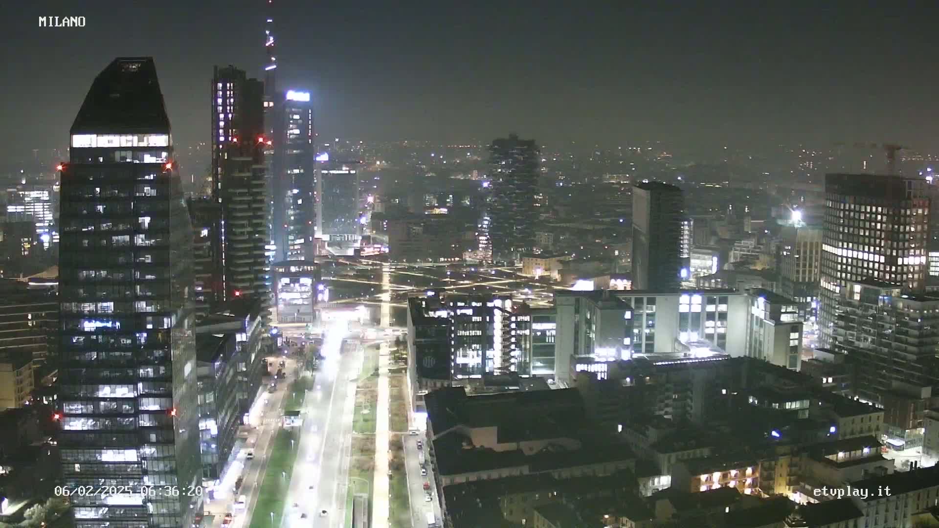 A nighttime, hazy view of a city skyline featuring numerous illuminated skyscrapers and streets.