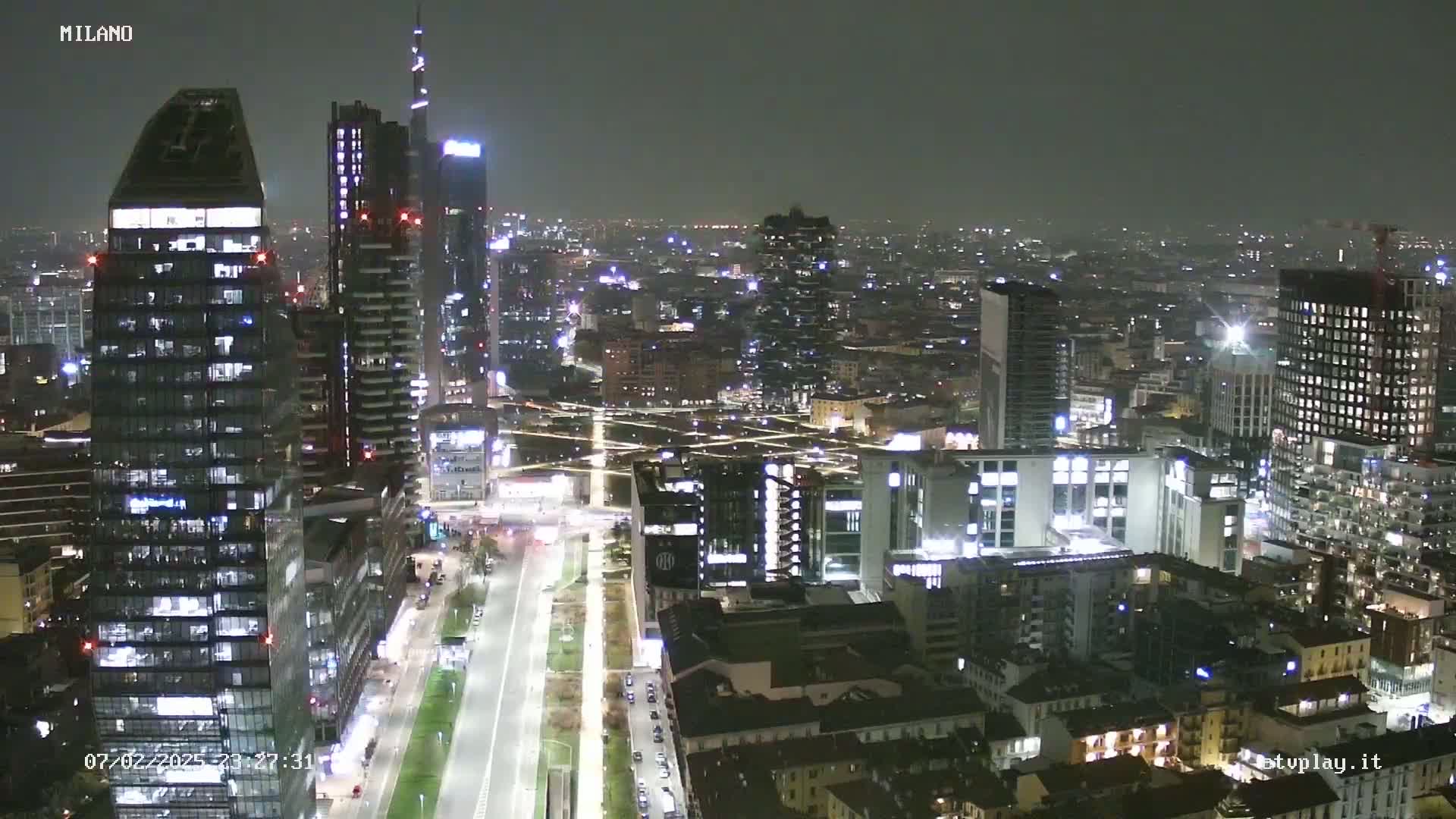 A nighttime aerial view of a city with numerous illuminated skyscrapers and buildings, under a clear, dark sky.