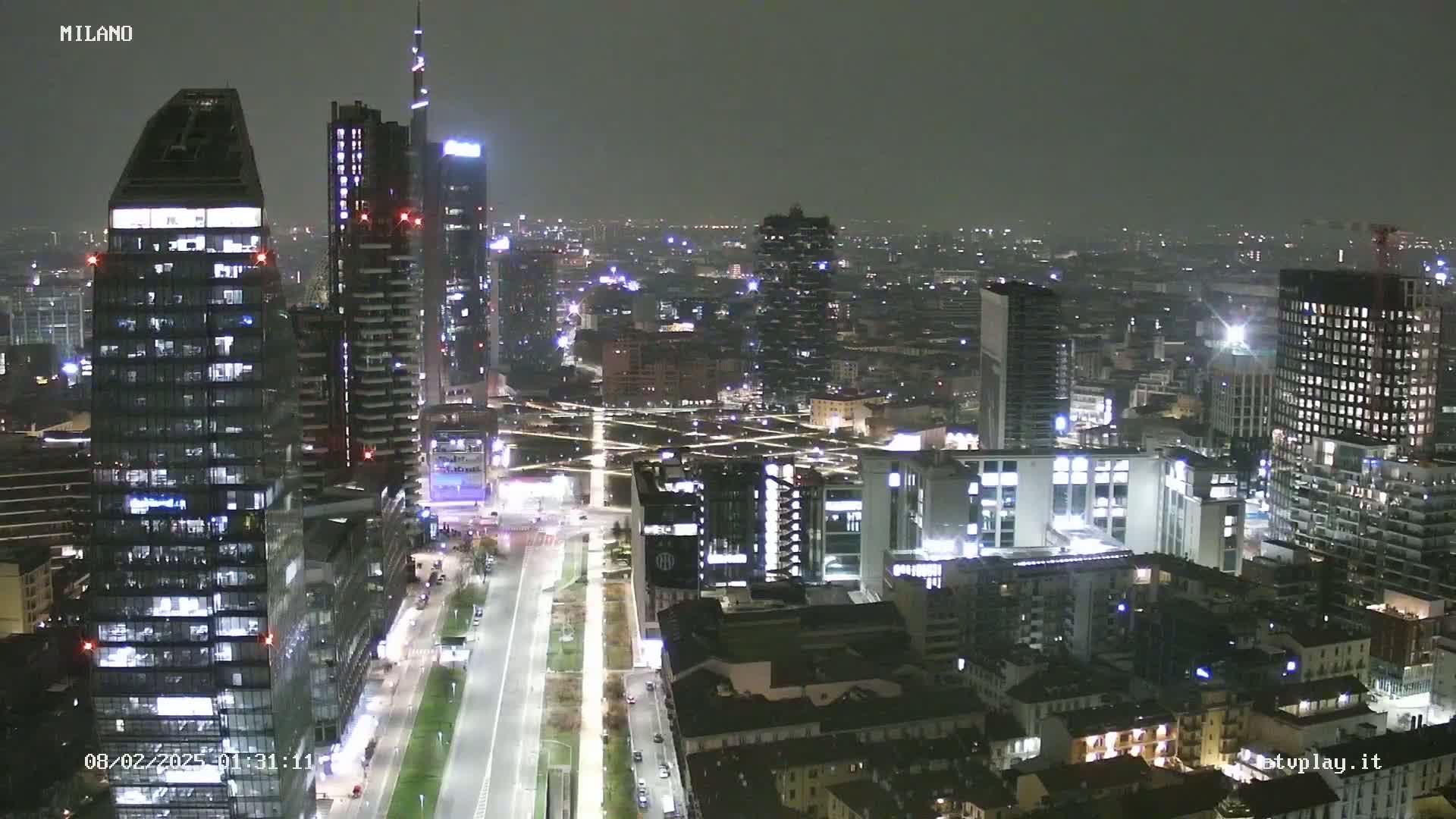 A nighttime aerial view of a city with many tall buildings illuminated, under a dark, possibly hazy sky.