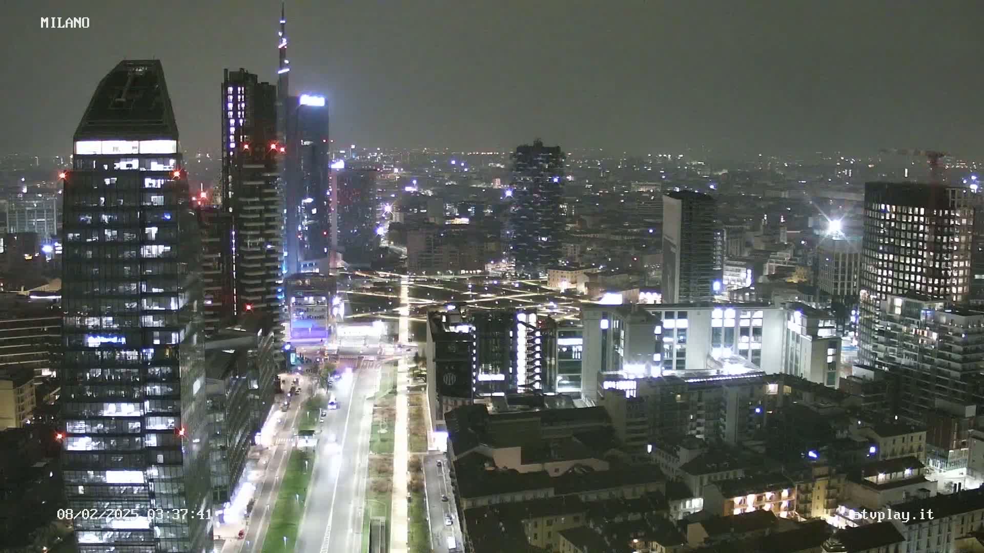 A nighttime aerial view of a city with many tall buildings illuminated, under a dark, overcast sky.