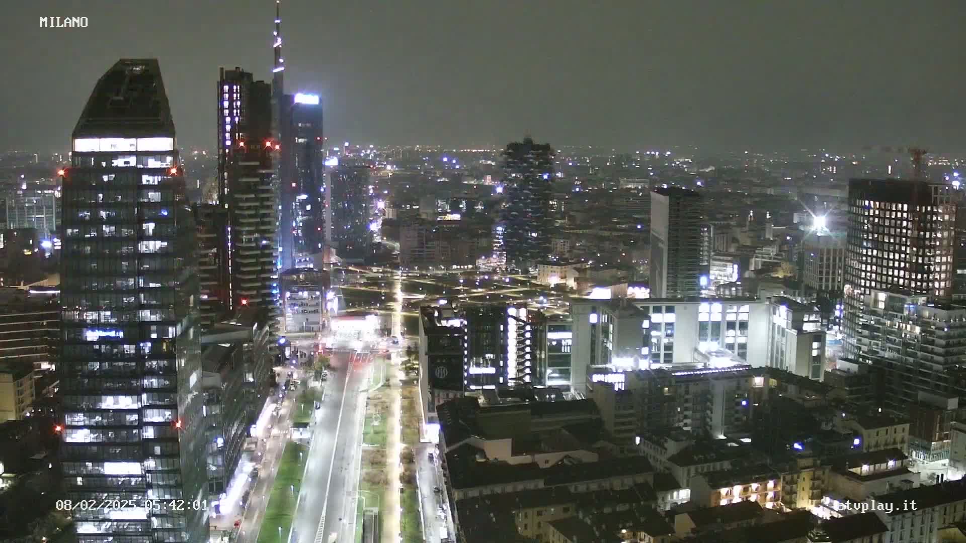 A nighttime aerial view of a city skyline with many tall buildings illuminated, under a cloudy sky.