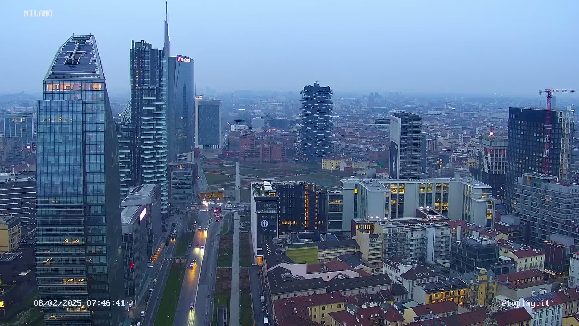 A hazy, overcast cityscape view of Milan, Italy, showing numerous modern skyscrapers and surrounding buildings along a wide road.