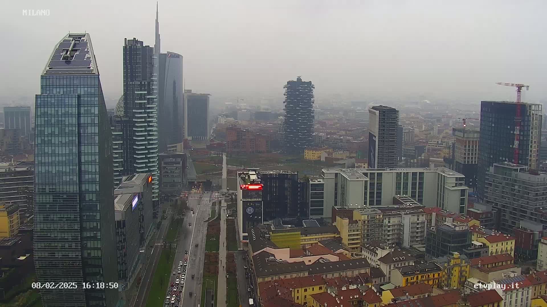 A hazy, overcast view of a dense cityscape featuring numerous skyscrapers and high-rise buildings, with a long road running through the center.