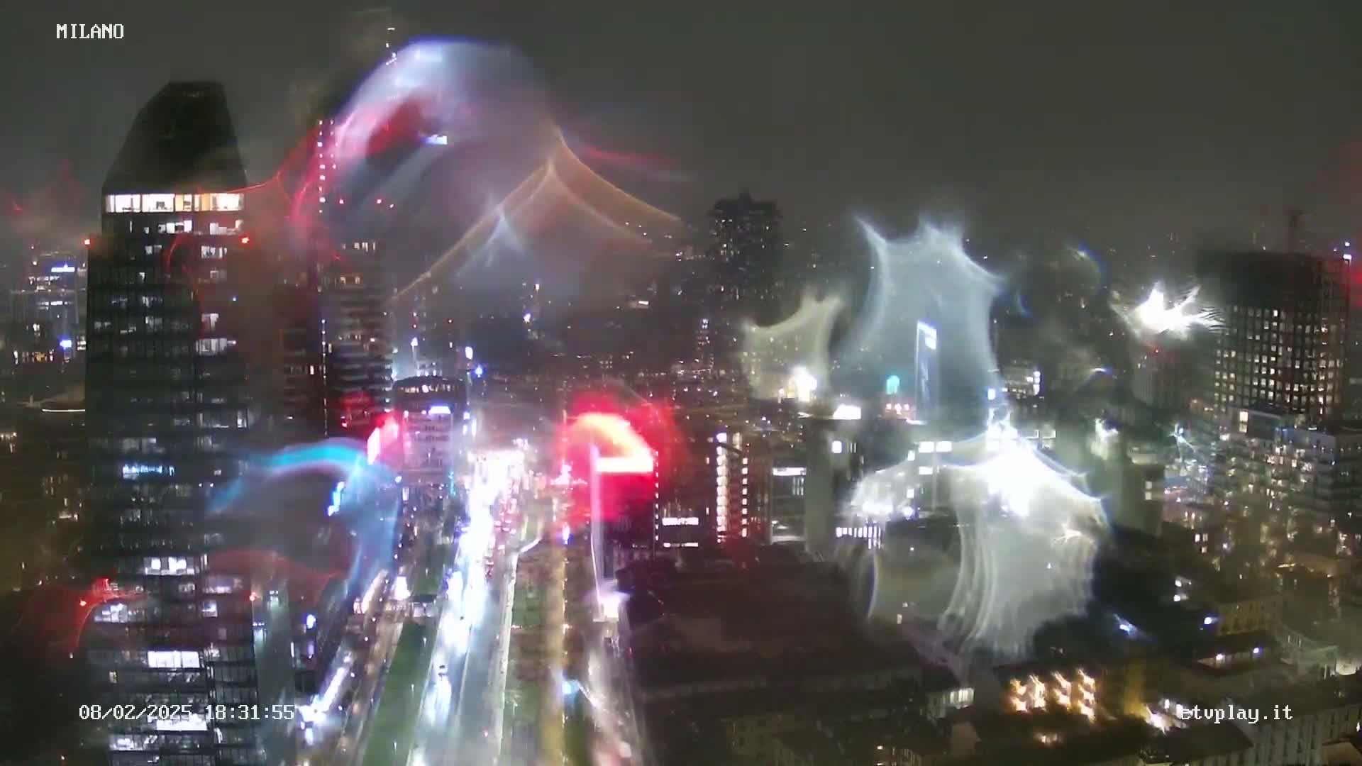 A nighttime, rain-streaked view of a city skyline with illuminated buildings and roadways, overlaid by streaks of light that appear to be caused by rain or reflections on a camera lens.
