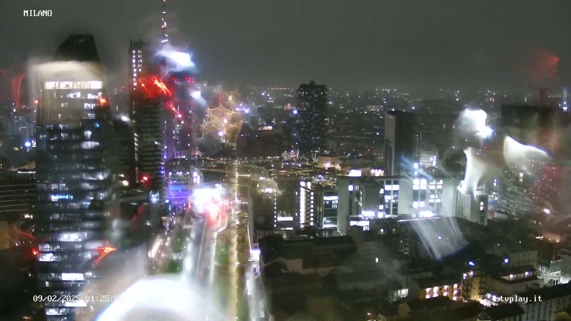 A nighttime view of a city skyline with tall buildings, illuminated streets, and what appears to be rain or light snow.
