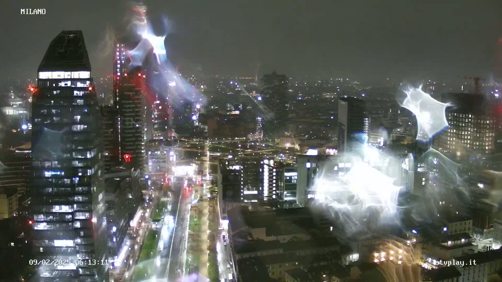 A nighttime, rain-streaked view of a city skyline showing illuminated buildings and streets.