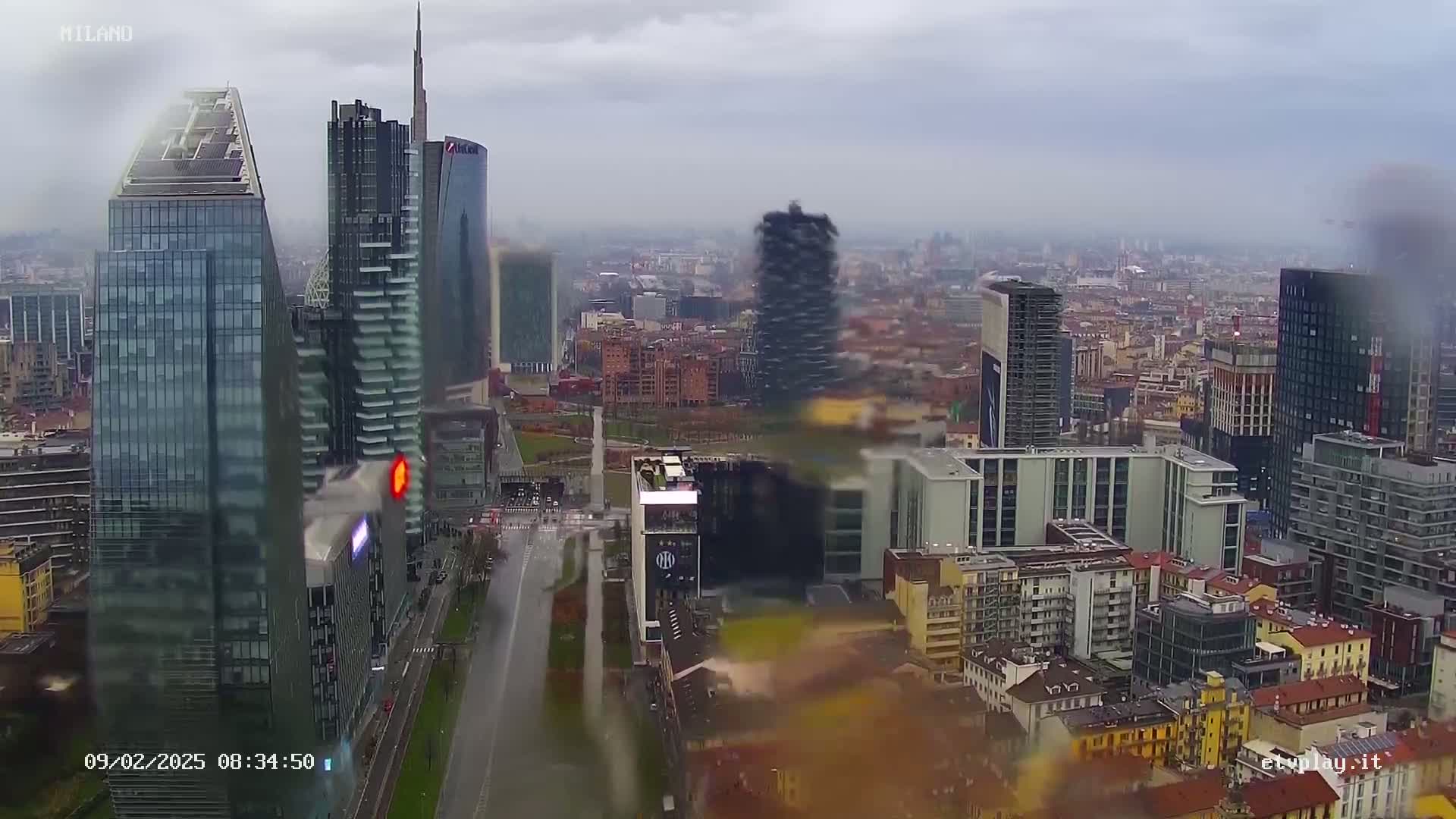 A rainy view of a cityscape featuring numerous skyscrapers and high-rise buildings, with a long road stretching through the foreground.