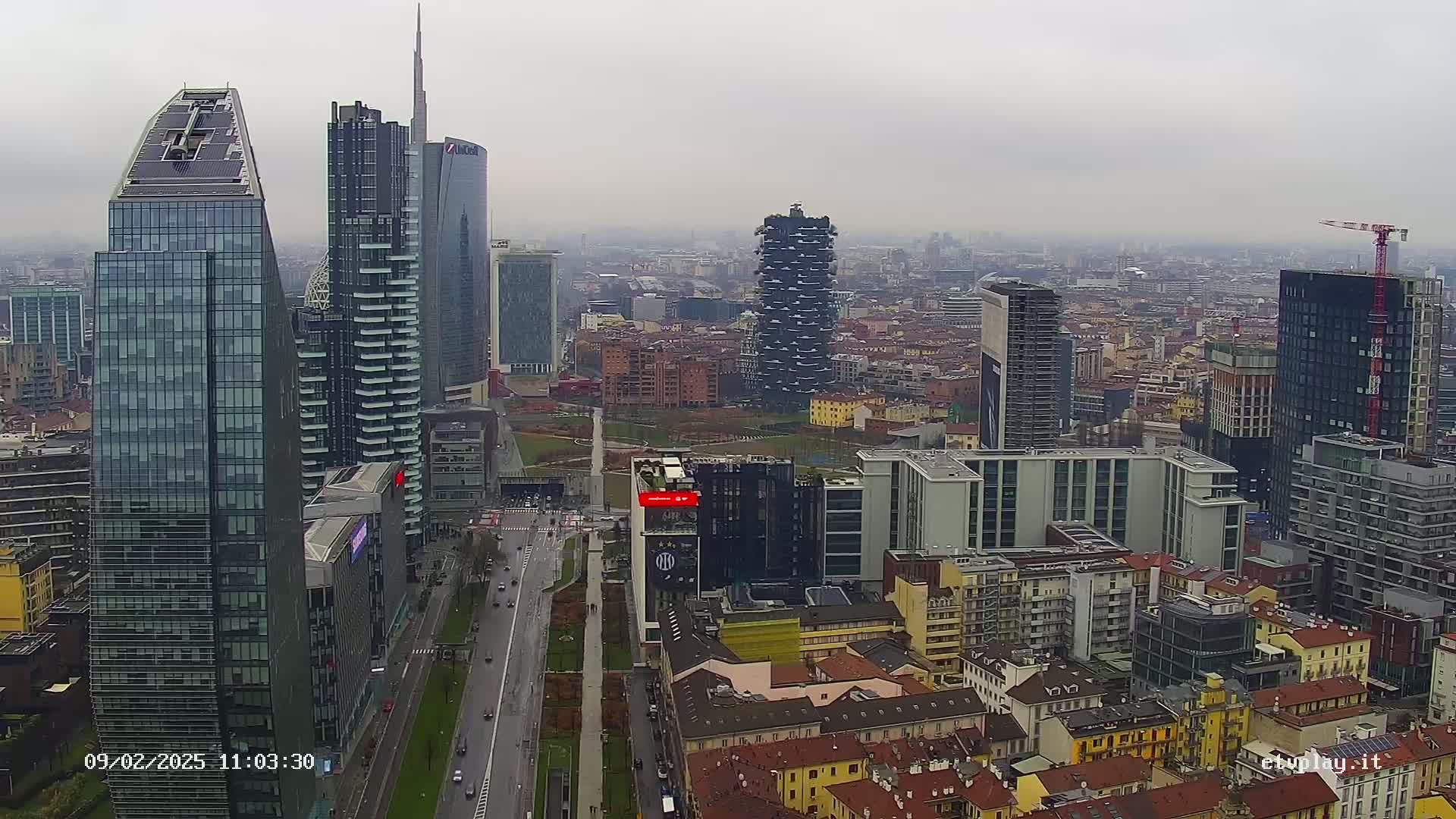 A high-angle, long shot of a city skyline on an overcast day, showing numerous skyscrapers and surrounding buildings, with a street visible below.