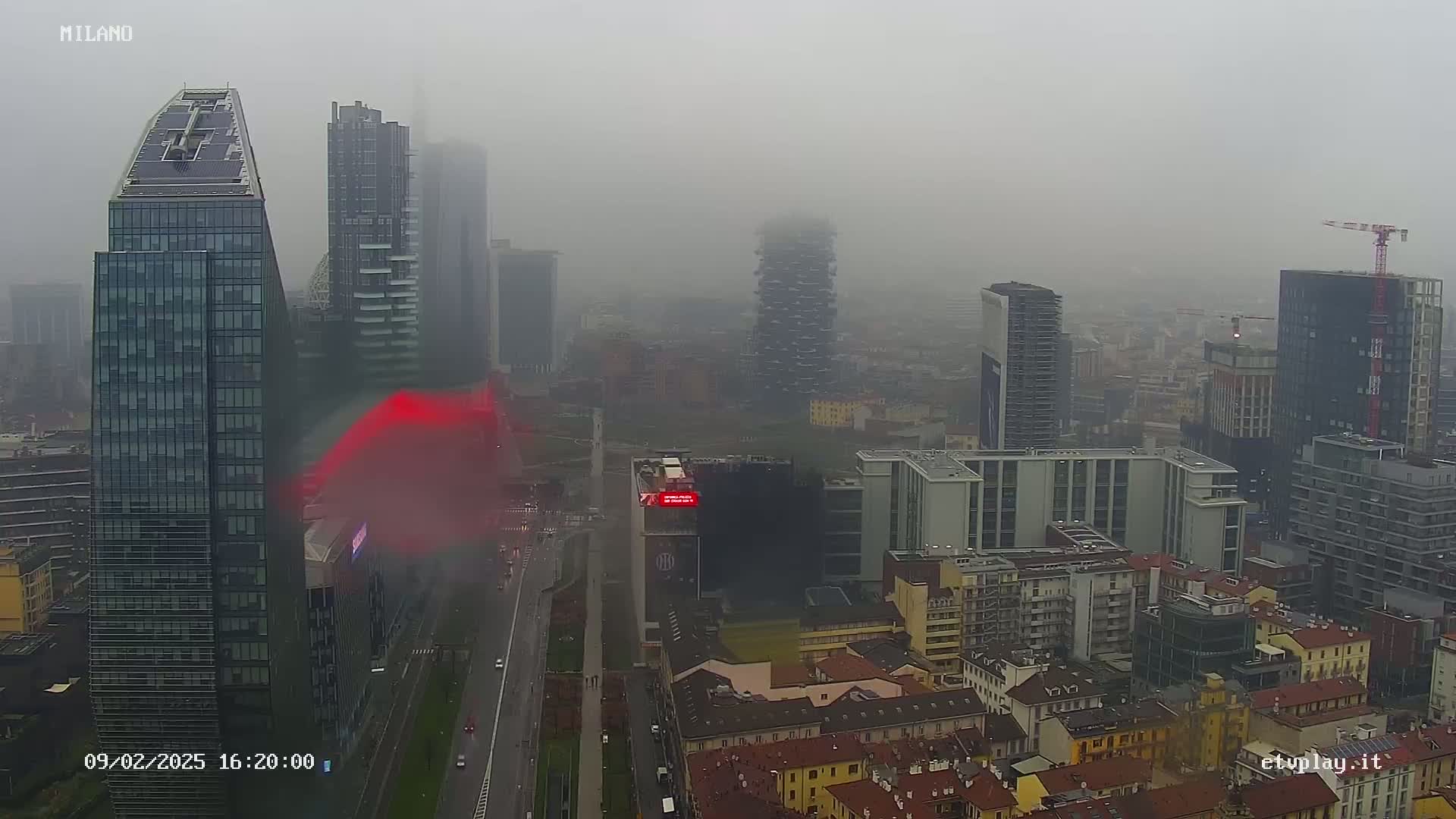 A foggy cityscape of many tall buildings, a road, and a construction crane, viewed from above.