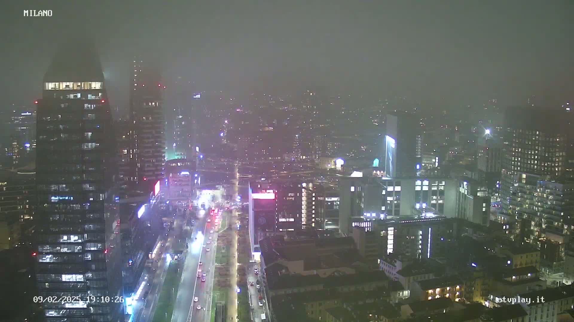 A nighttime, foggy view of a city with numerous tall buildings and a highway with traffic visible below.