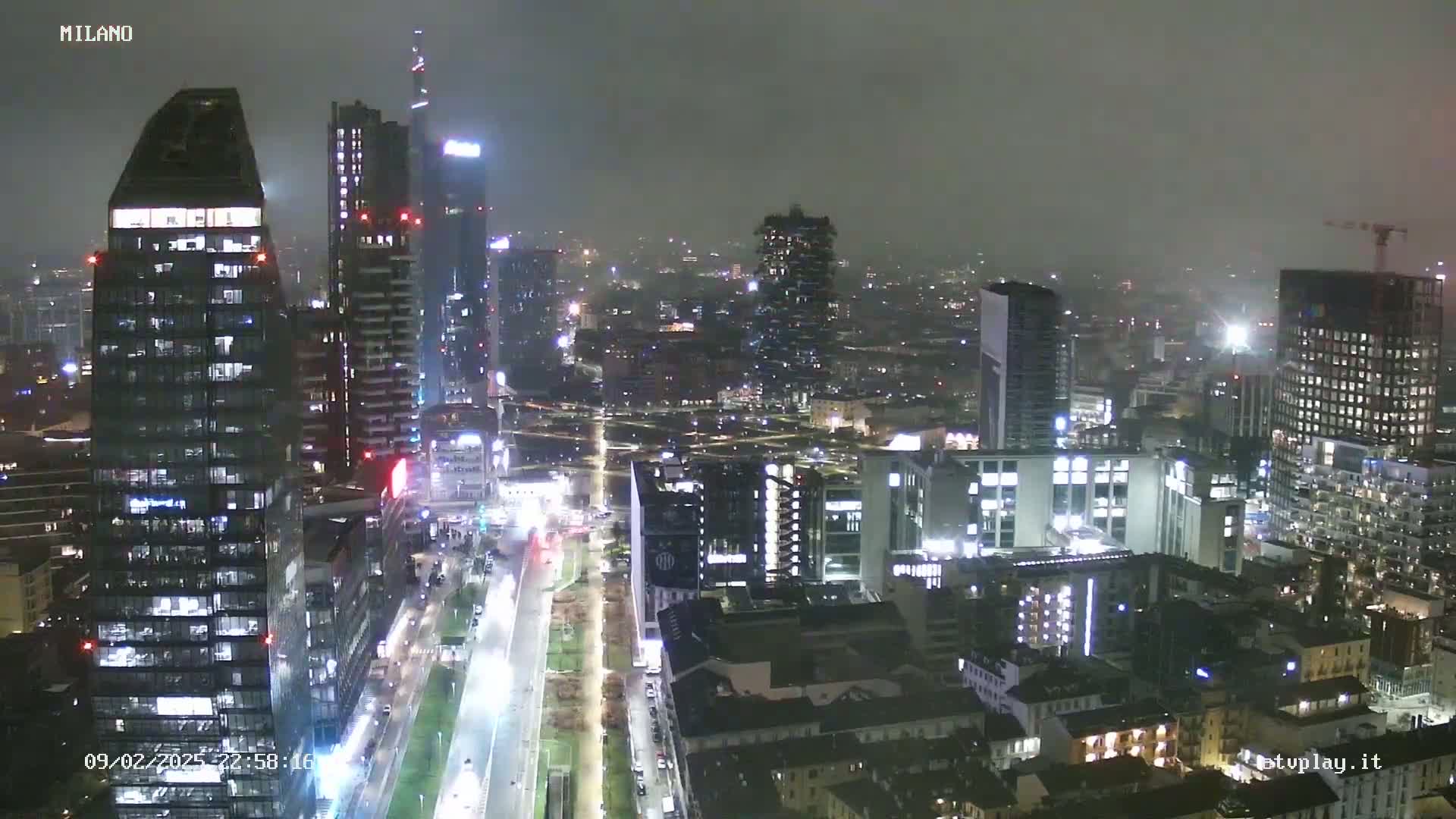 A nighttime aerial view of a city, possibly Milan, shows numerous illuminated skyscrapers and buildings under an overcast sky.