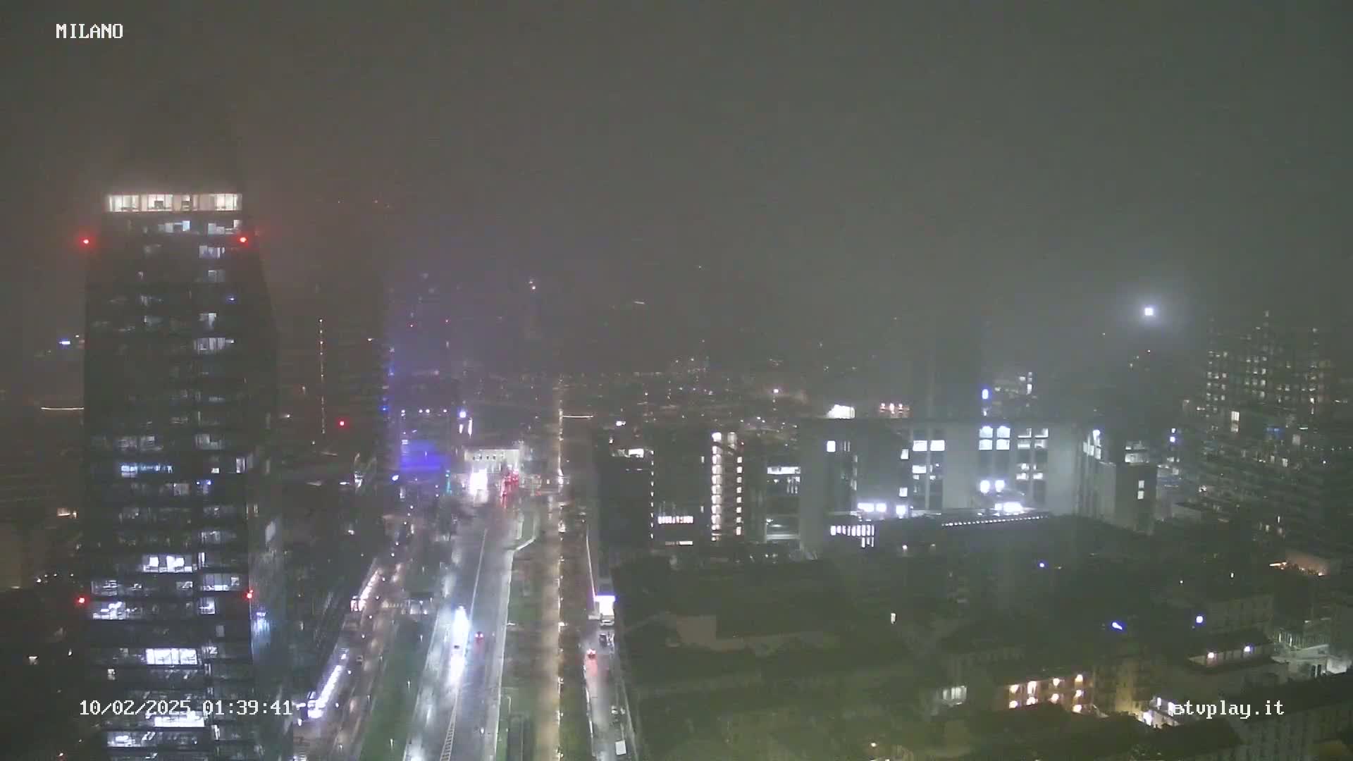 A nighttime, high-angle view of a city shrouded in heavy fog or mist, showing illuminated buildings and a long, wet road.