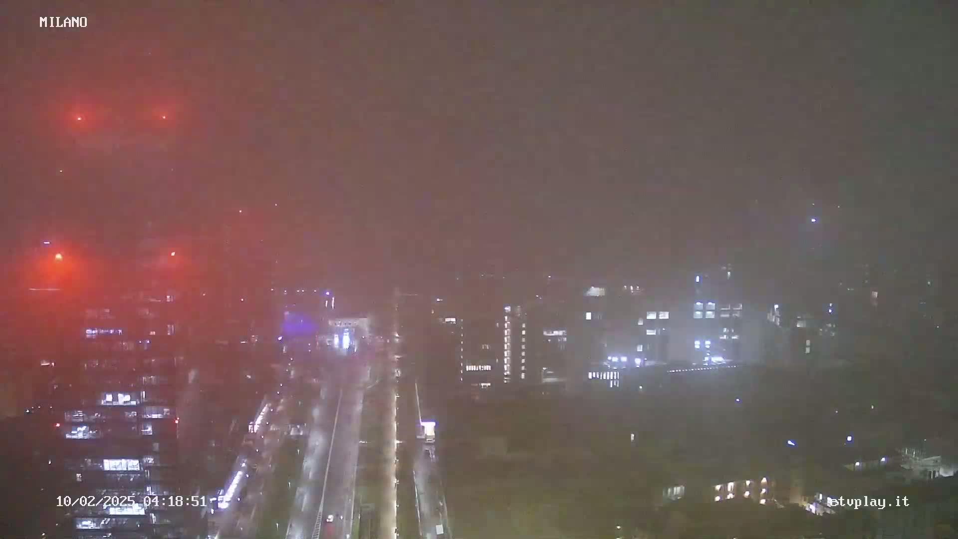 A nighttime, hazy view of city buildings and a road, with faint lights visible through the atmospheric conditions.