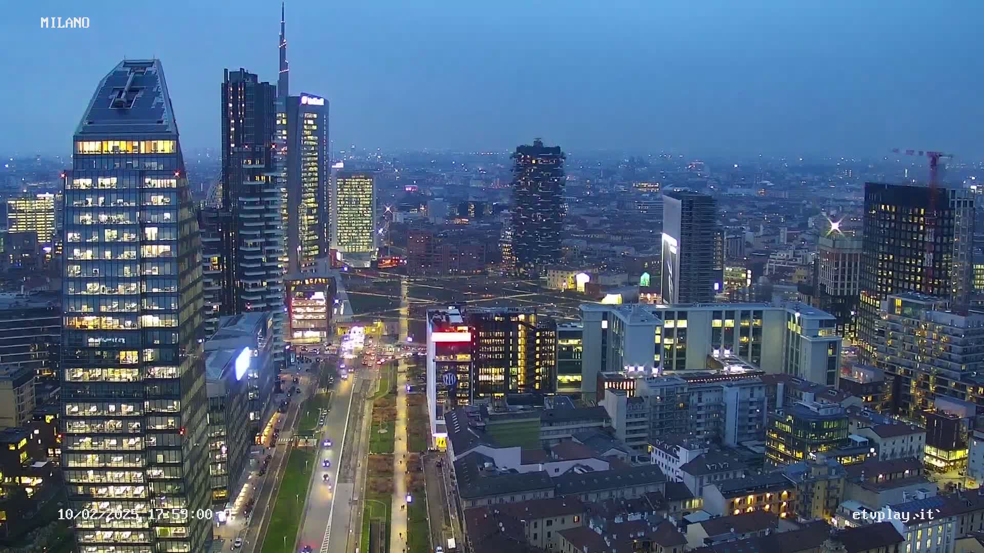 A city skyline at twilight, showing numerous illuminated skyscrapers and buildings, with a long, tree-lined avenue in the foreground.
