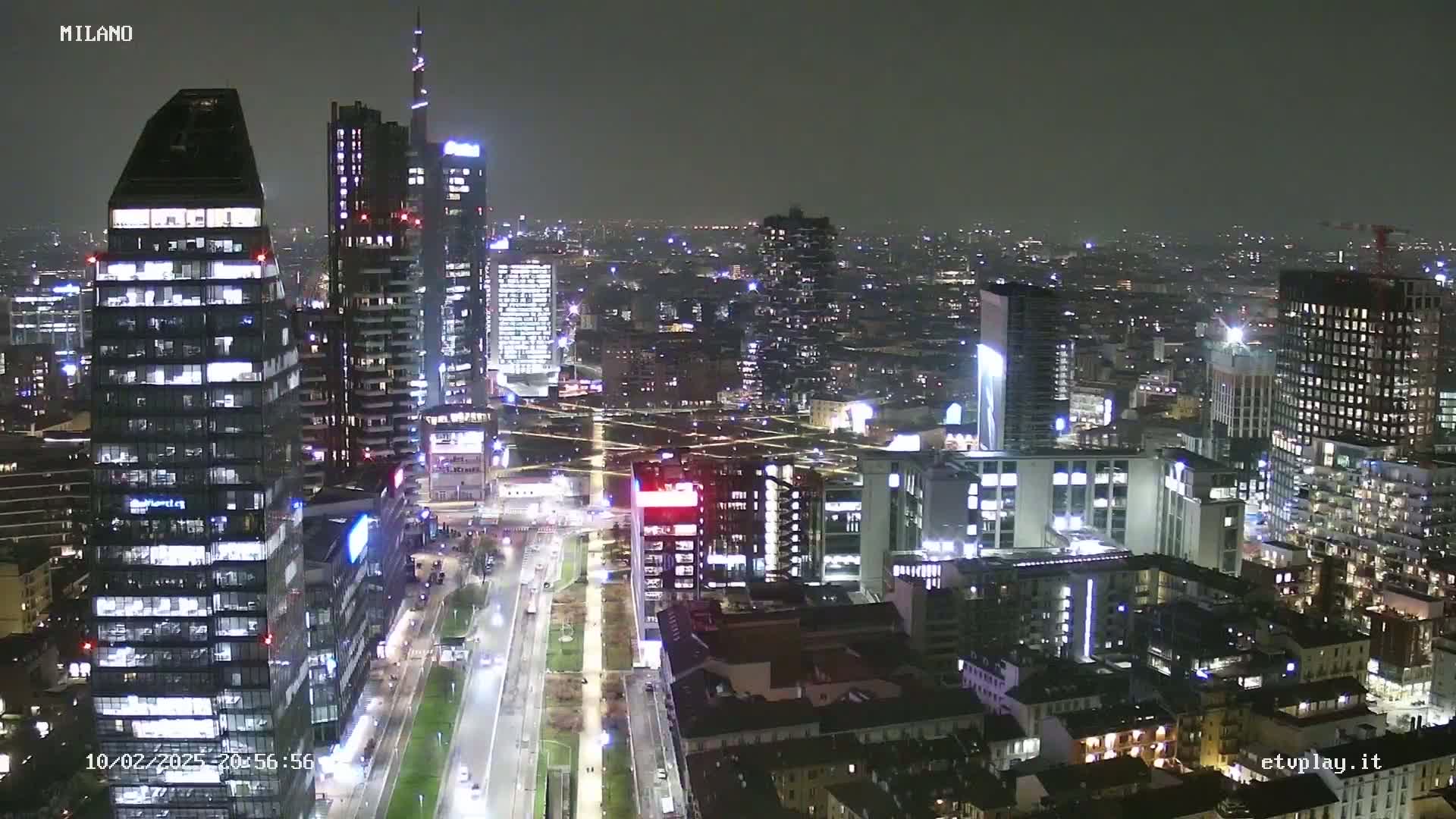 A nighttime aerial view of a city's illuminated skyline, showing many tall buildings and streets with vehicle lights, under a dark, clear sky.