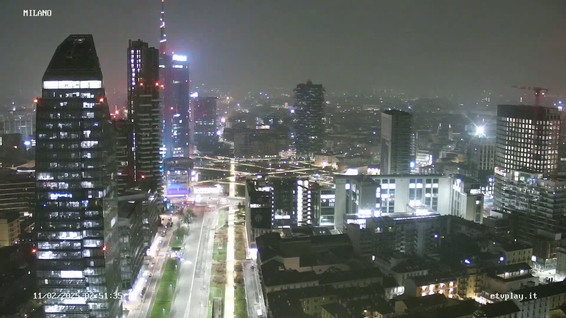 A nighttime aerial view of a city skyline with many illuminated skyscrapers under hazy conditions.