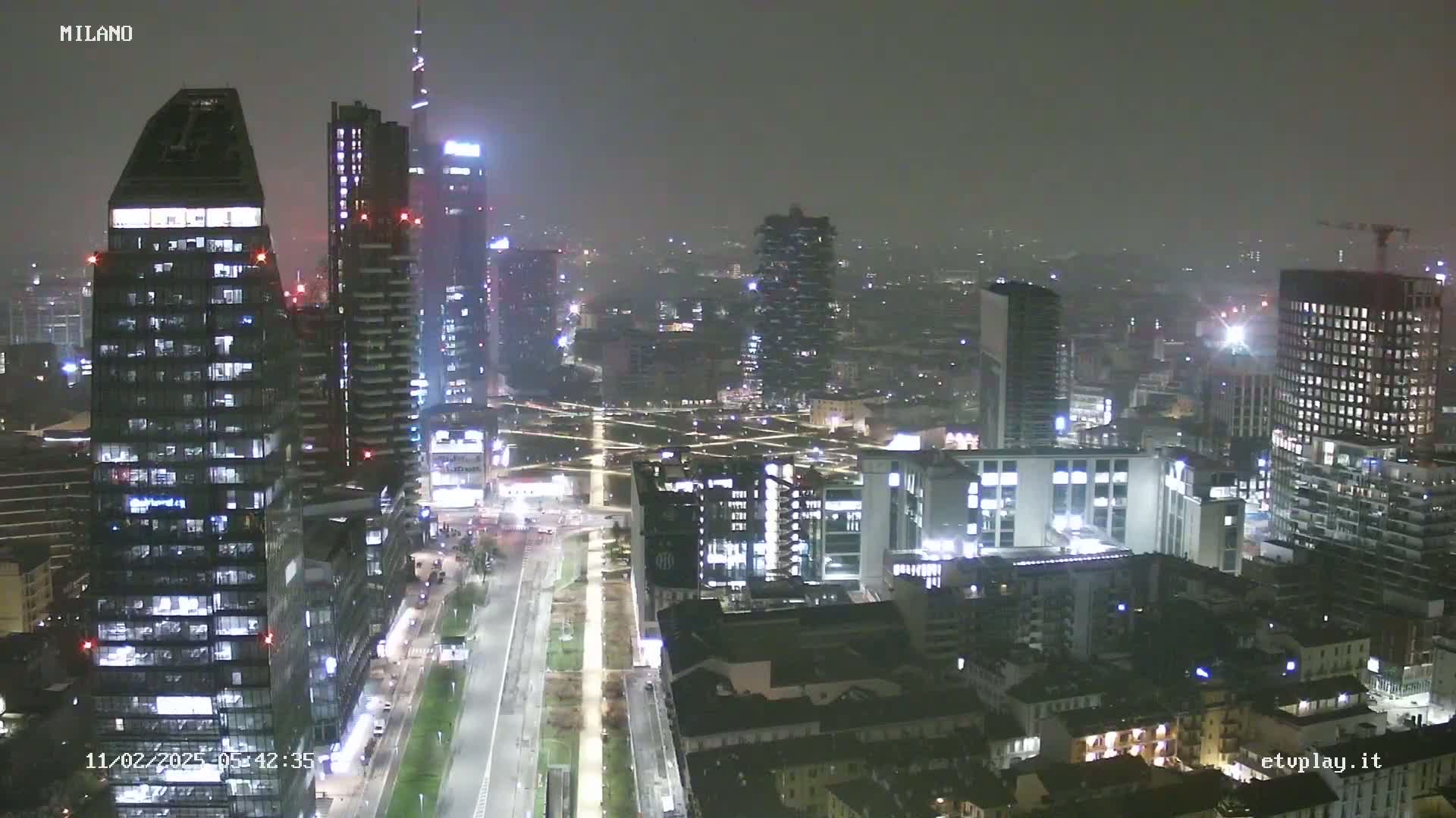 A nighttime aerial view of a city, possibly Milan, shows numerous illuminated skyscrapers and buildings under hazy or foggy conditions.