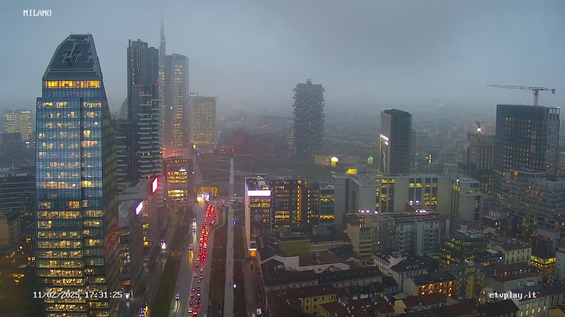 A nighttime view of a city skyline with many tall buildings and a busy road underneath, under an overcast, rainy sky.