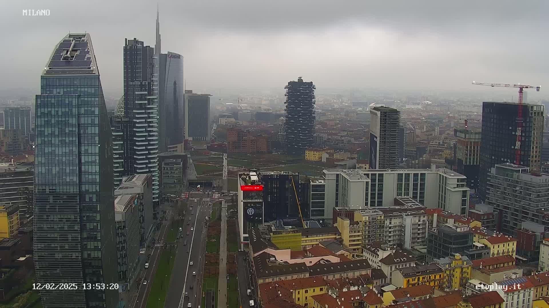 A high-angle, long shot of a city skyline on an overcast day, showing numerous modern skyscrapers and surrounding buildings, with a roadway visible below.