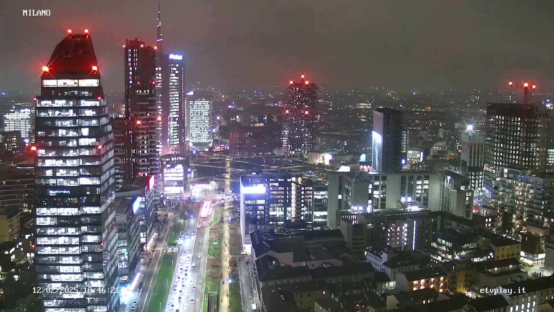A nighttime aerial view of a city's skyline showing many illuminated skyscrapers and a roadway with traffic under hazy or foggy conditions.