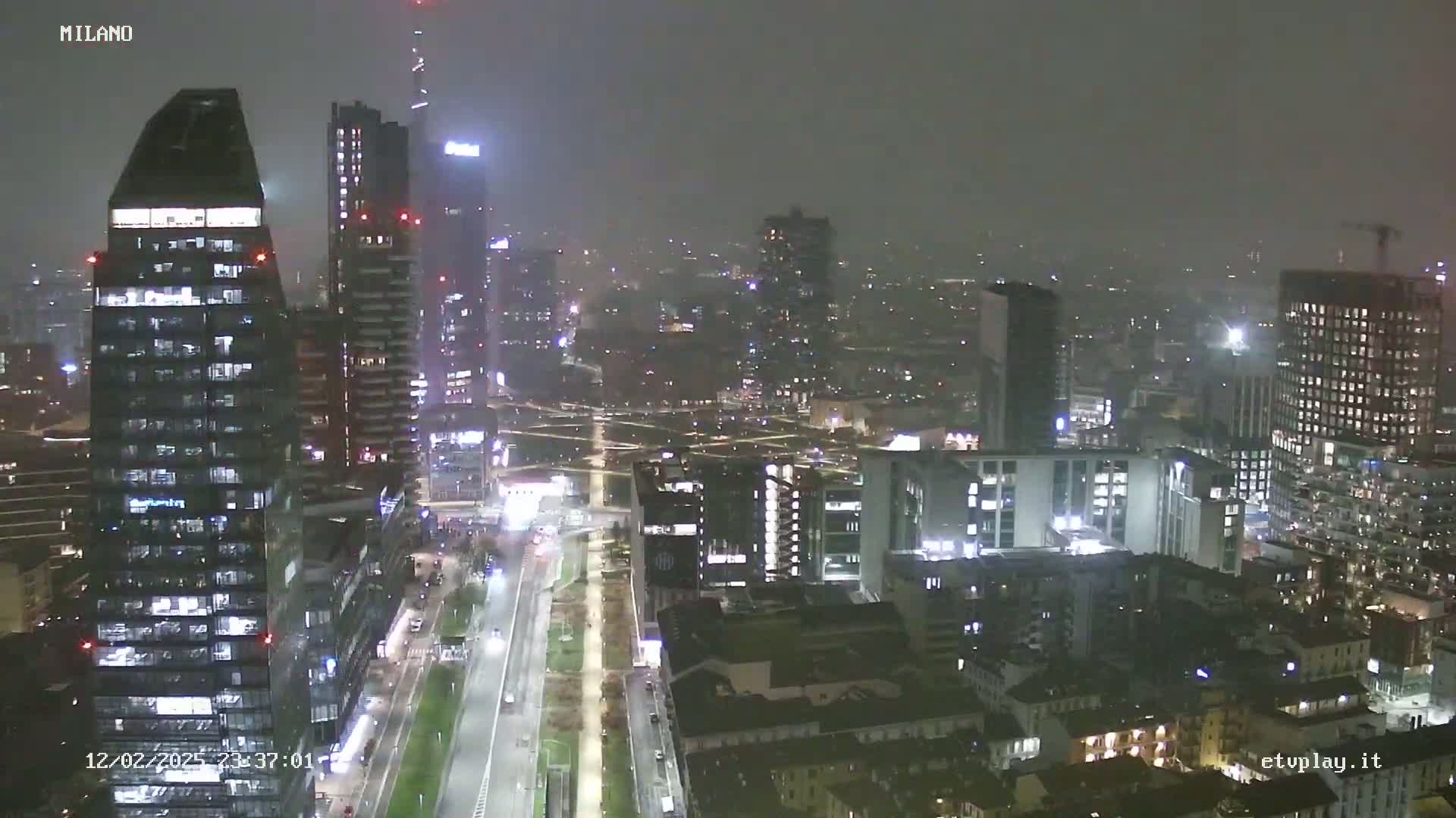 A nighttime, hazy view of a city skyline showing many tall buildings with lights on, and a road below.